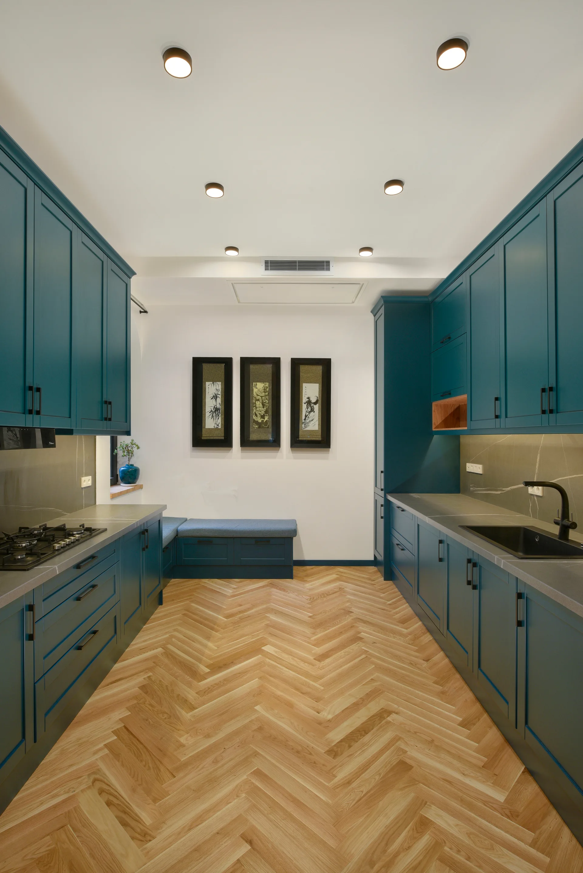 A galley kitchen featuring deep teal shaker cabinets, light wood herringbone flooring, grey stone countertops, and a built-in corner bench at the far end.