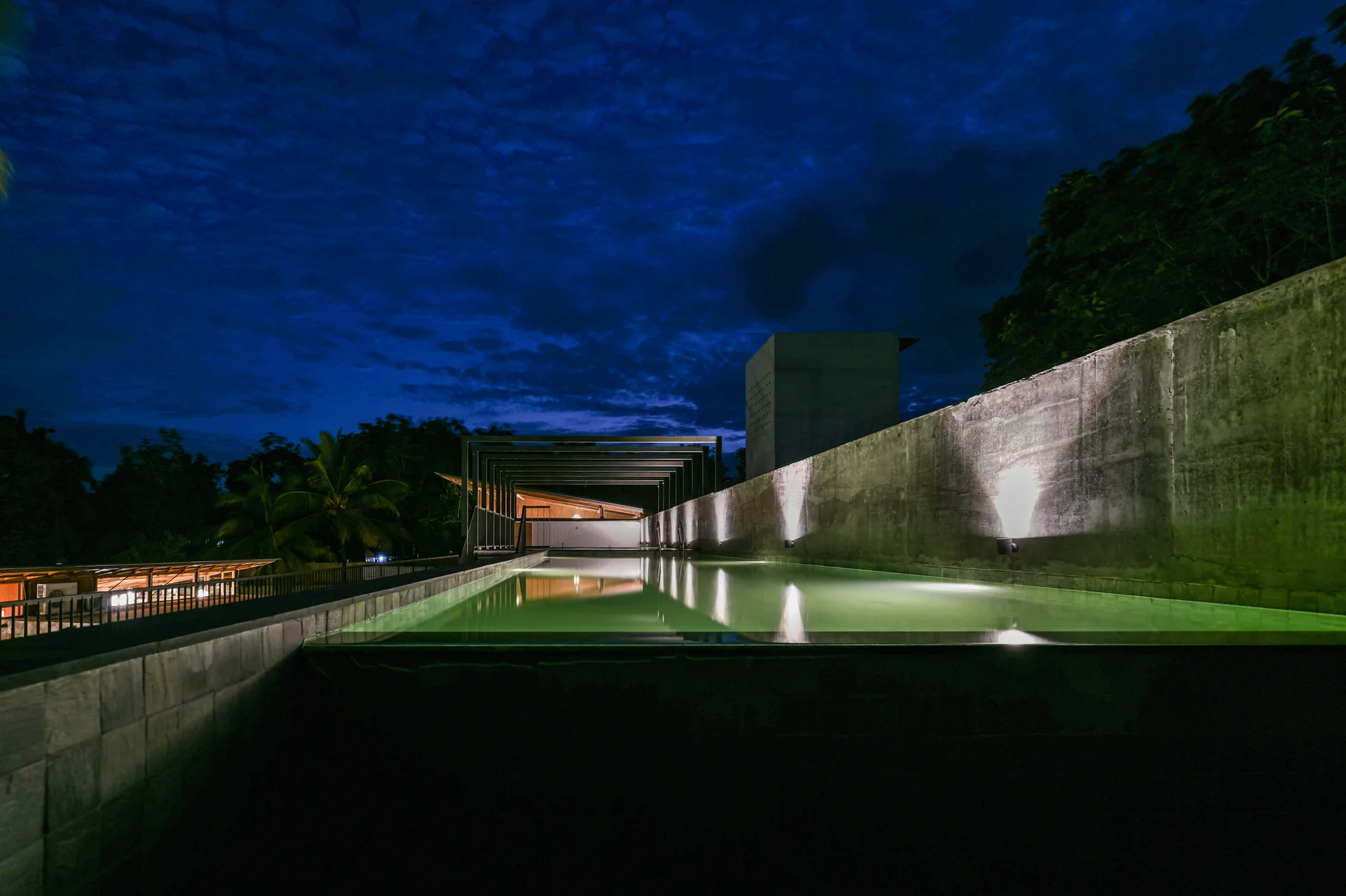 An illuminated outdoor lap pool runs alongside a textured concrete wall with warm uplighting under a dark twilight sky.