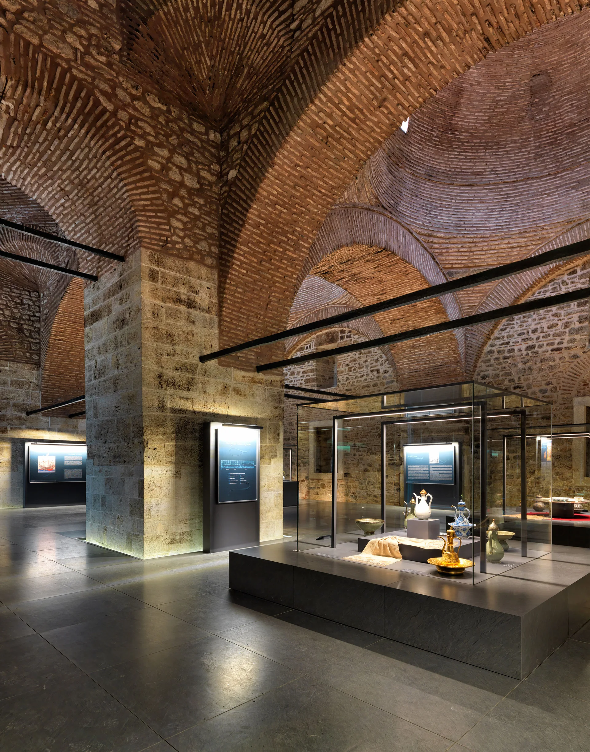 Modern glass exhibition cases displaying artifacts stand under historic vaulted brick ceilings and massive stone pillars on a dark tile floor.