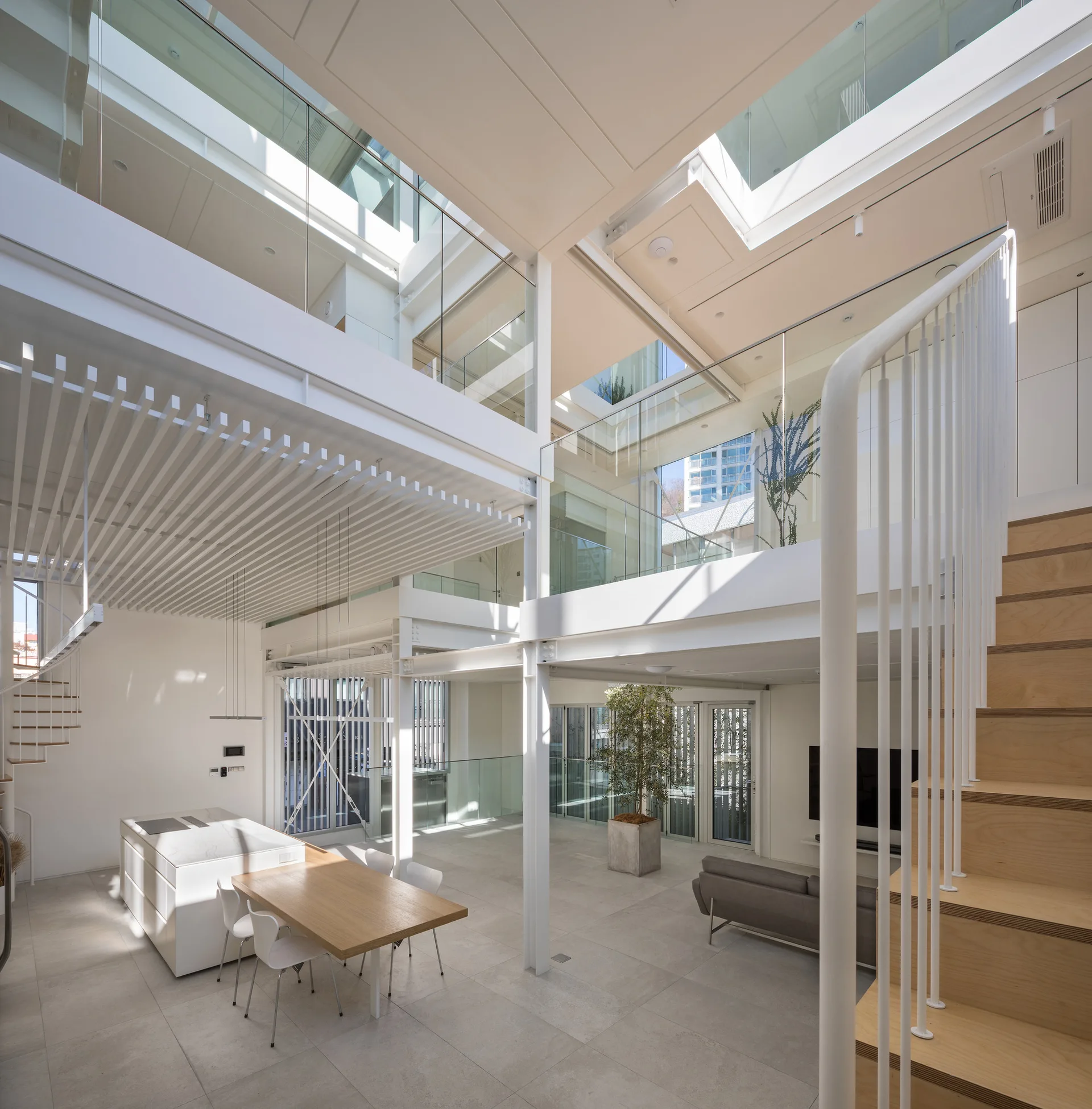 A modern multi-level living space with a white kitchen island, wooden dining table, staircase, and glass-railed mezzanines under skylights.
