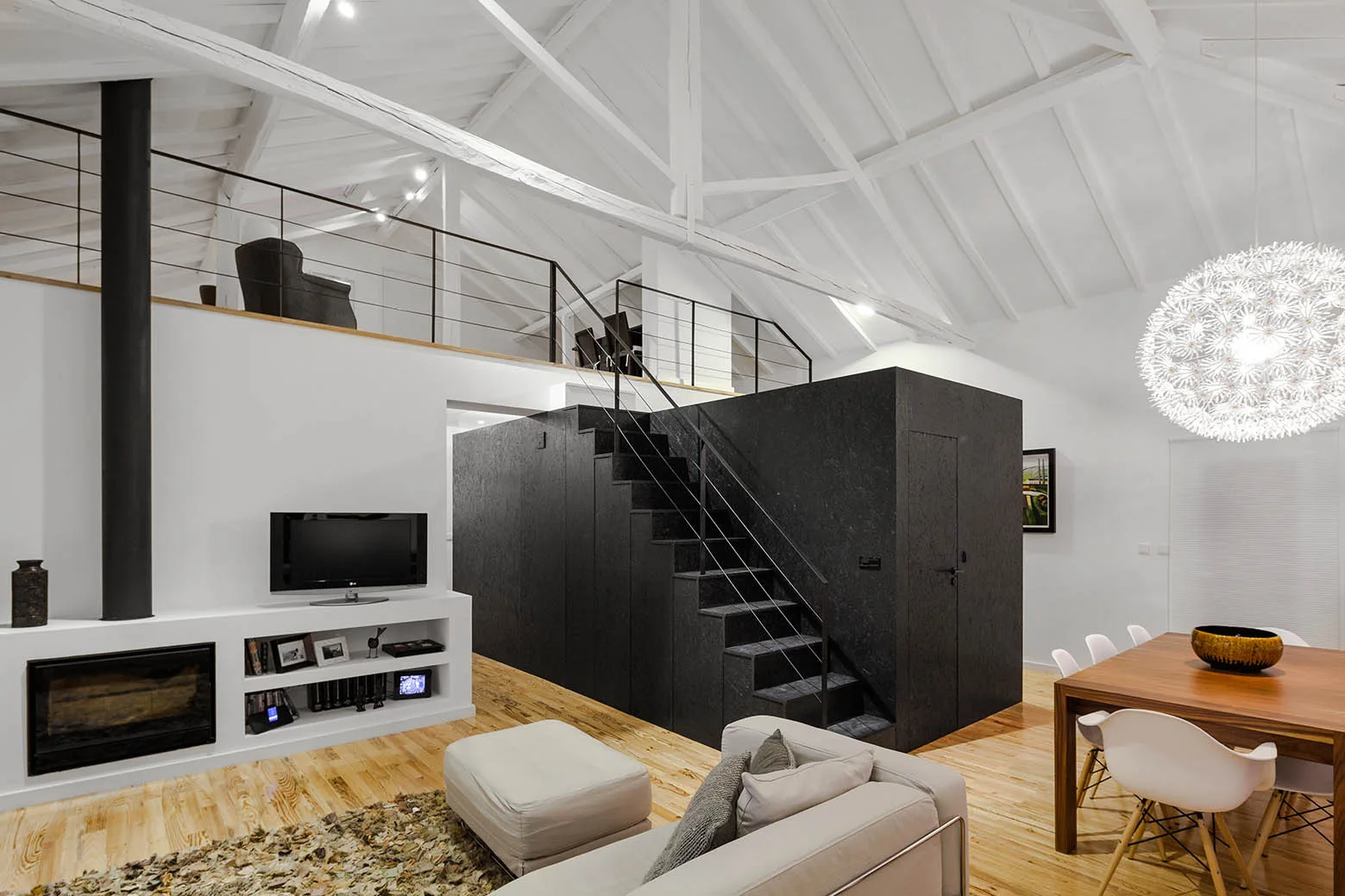 A modern loft featuring a central black staircase block, a dining area, and a white vaulted ceiling with exposed wooden trusses.