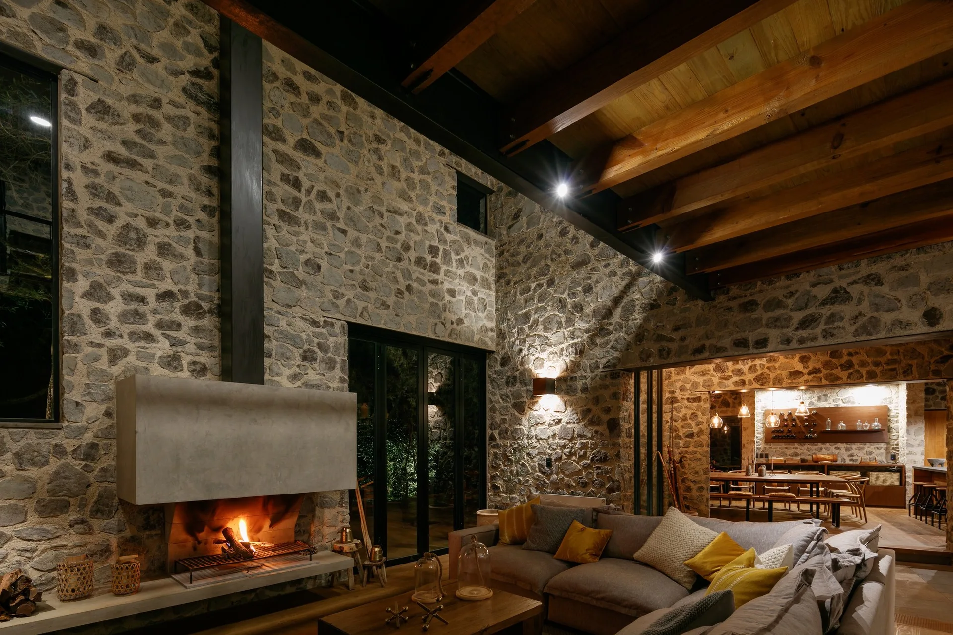 A rustic living room features a grey sectional sofa, an active concrete fireplace, and high walls made of exposed rough stone beneath wood ceiling beams.