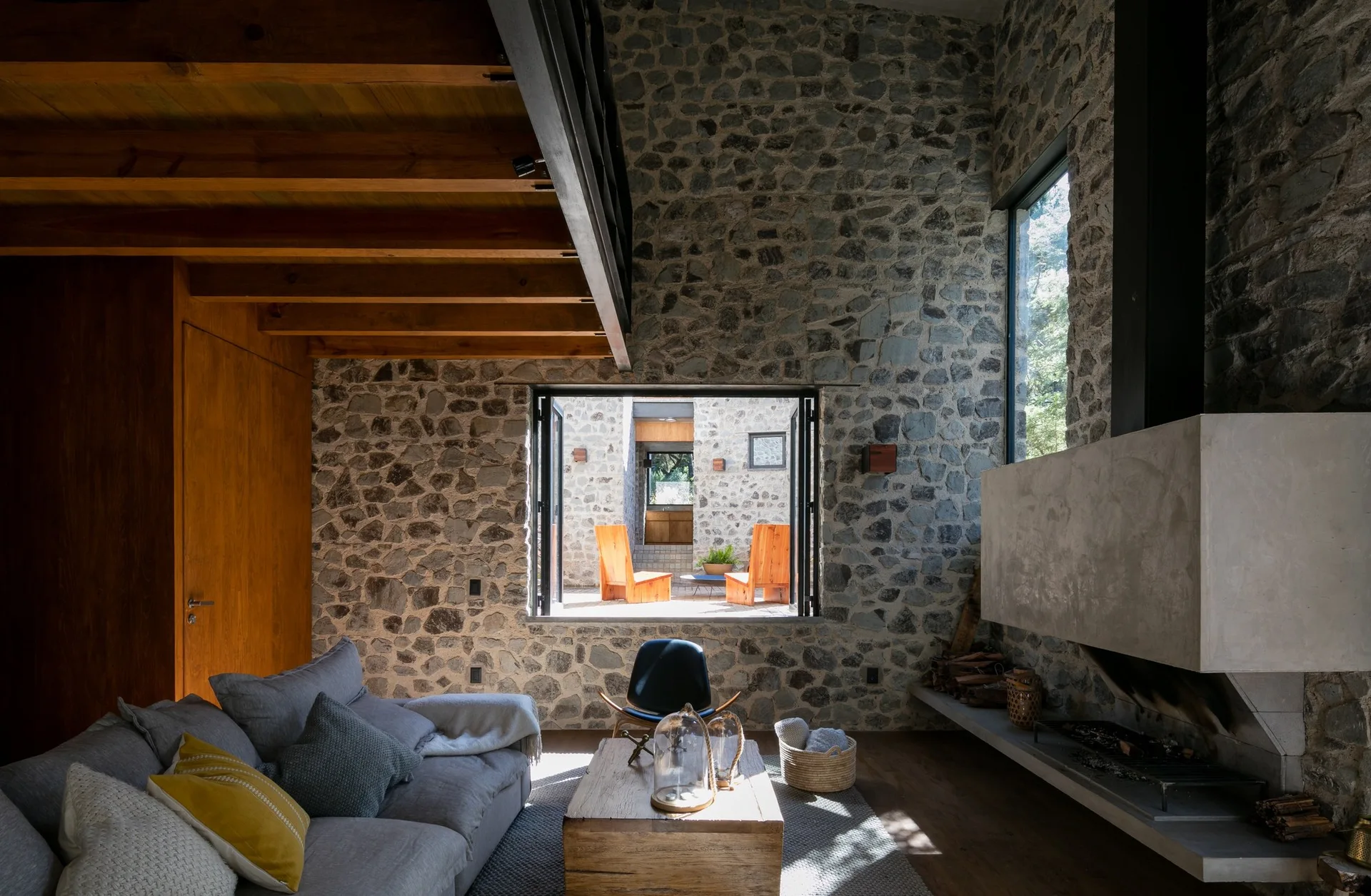 A rustic living room features a grey sofa, a wooden coffee table, an exposed stone wall, and a large window looking out onto a sunny courtyard.