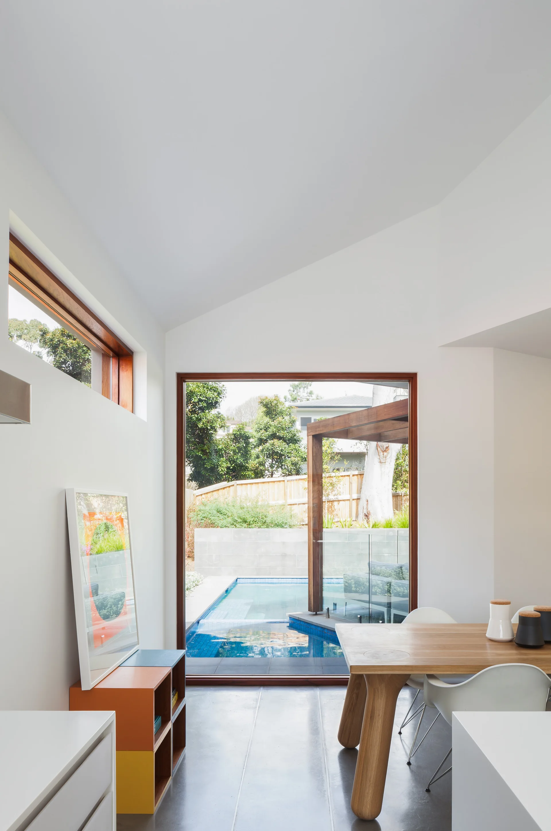 A bright dining area featuring a wooden table, white chairs, and a large window looking out to a blue backyard swimming pool.