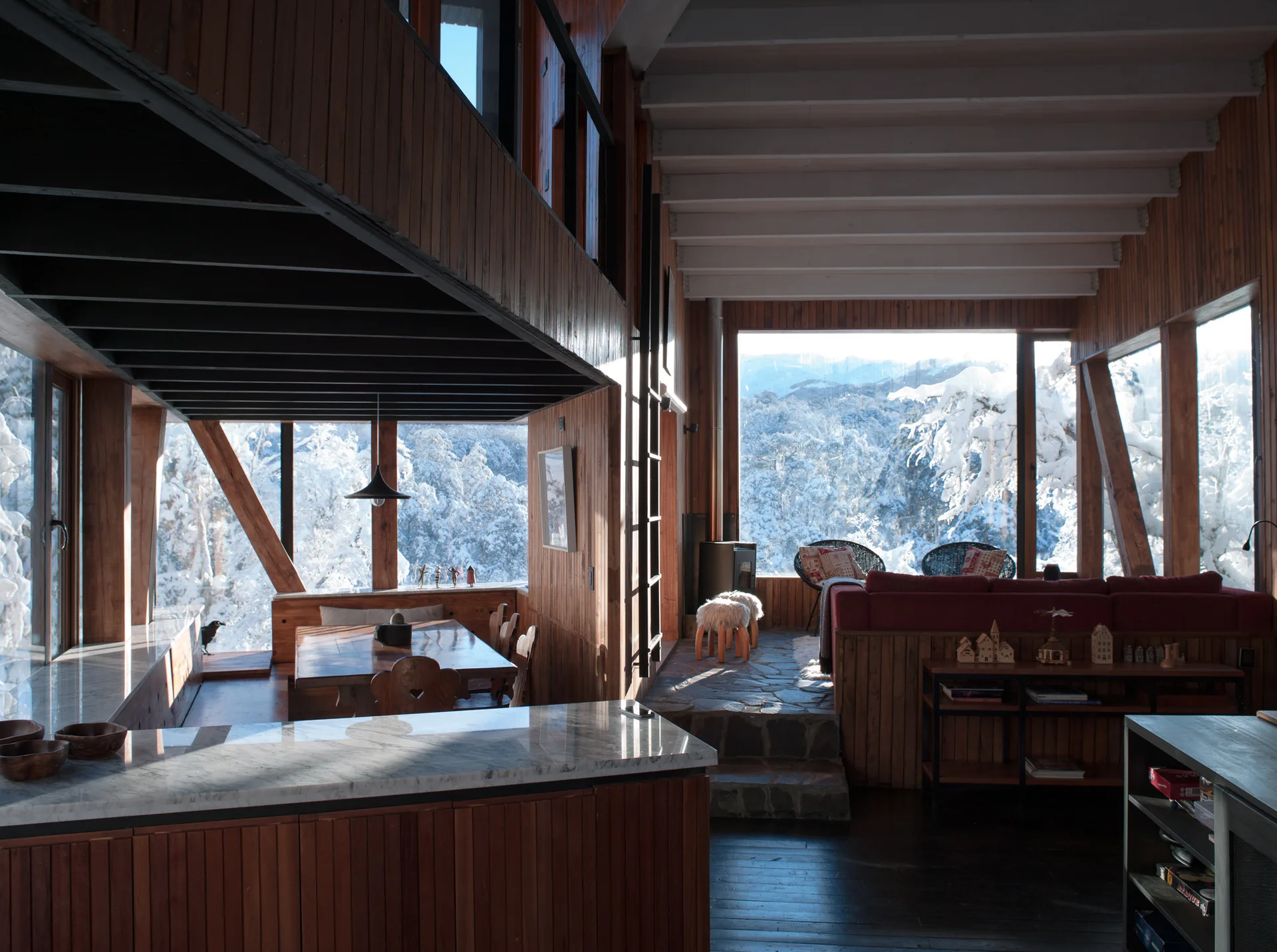 A rustic wood-paneled cabin interior featuring a split-level living area, dining table, marble kitchen counter, and large windows overlooking a snowy forest.