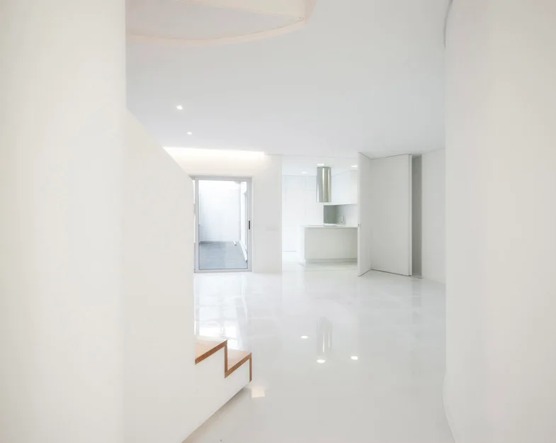 A minimalist all-white open interior with highly reflective glossy floors, curved walls, and wood-tread stairs in the foreground.