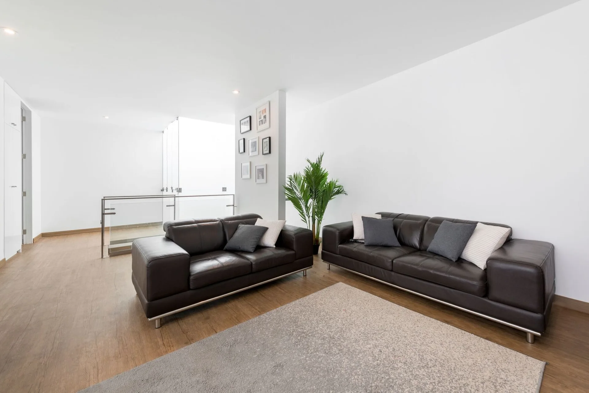 Two dark brown leather sofas face each other on a gray rug in a bright room with white walls, a glass railing, and wood floors.