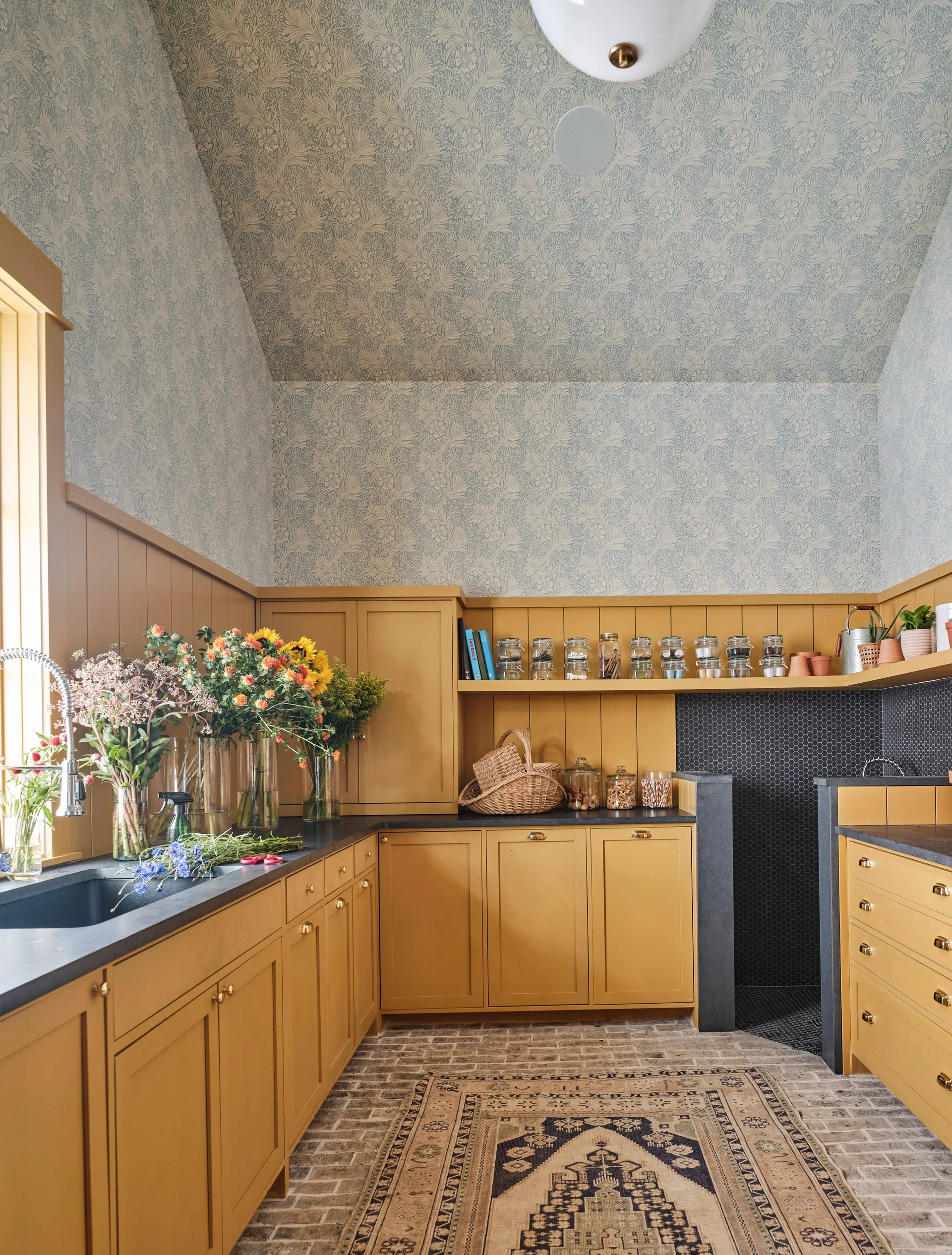 A utility room features mustard yellow cabinets, dark countertops, blue floral wallpaper on the walls and ceiling, and a brick floor with a vintage rug.