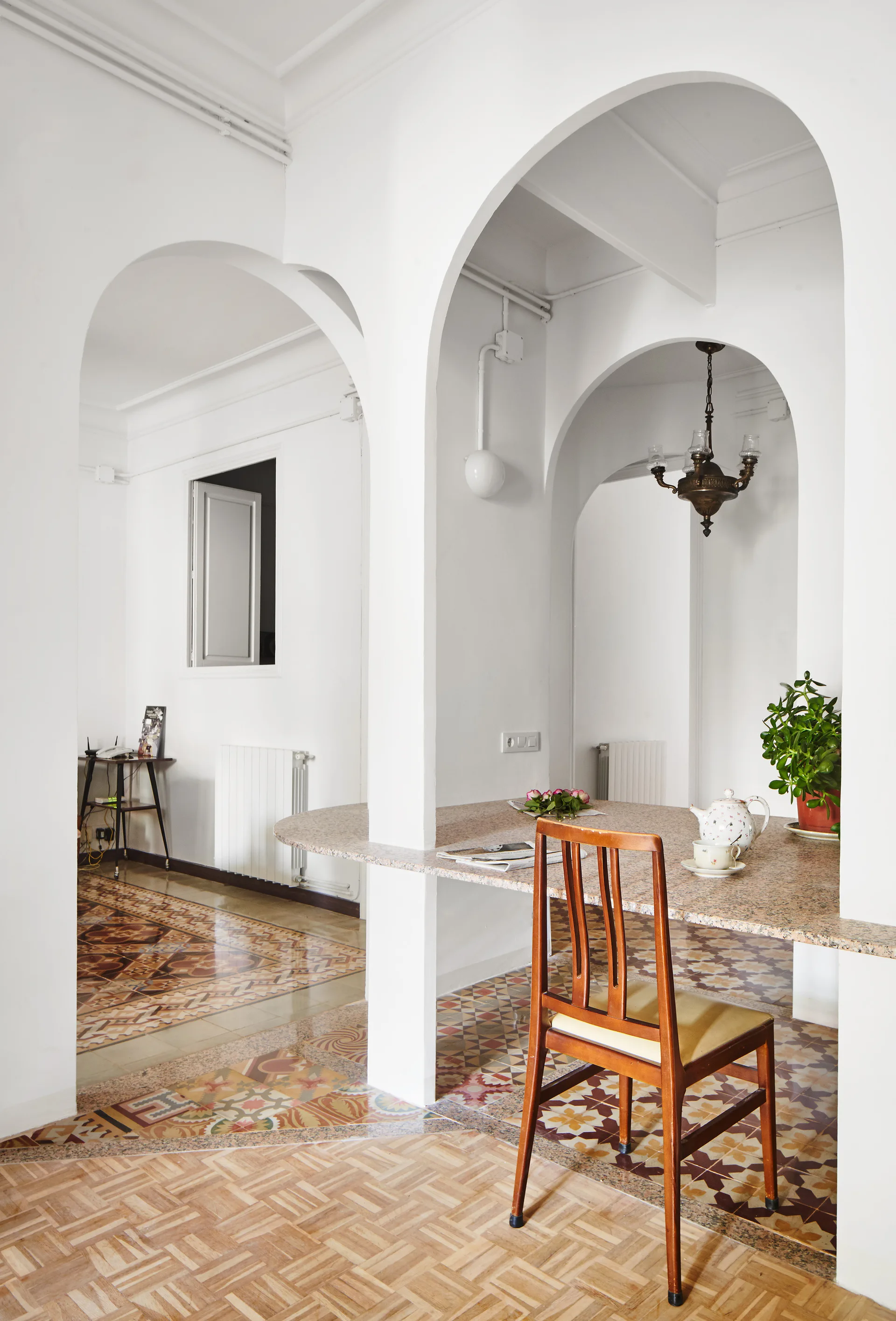 A dining area with a stone-topped table built into white structural arches, featuring a wooden chair and decorative mosaic floor tiles.