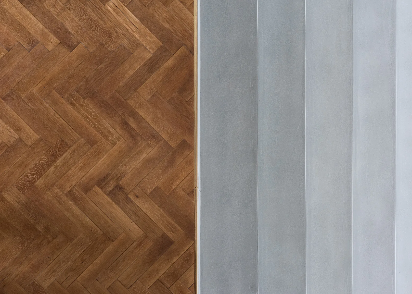 A top-down view of a flooring transition between herringbone wood and smooth grey concrete, separated by a thin gold metal strip.