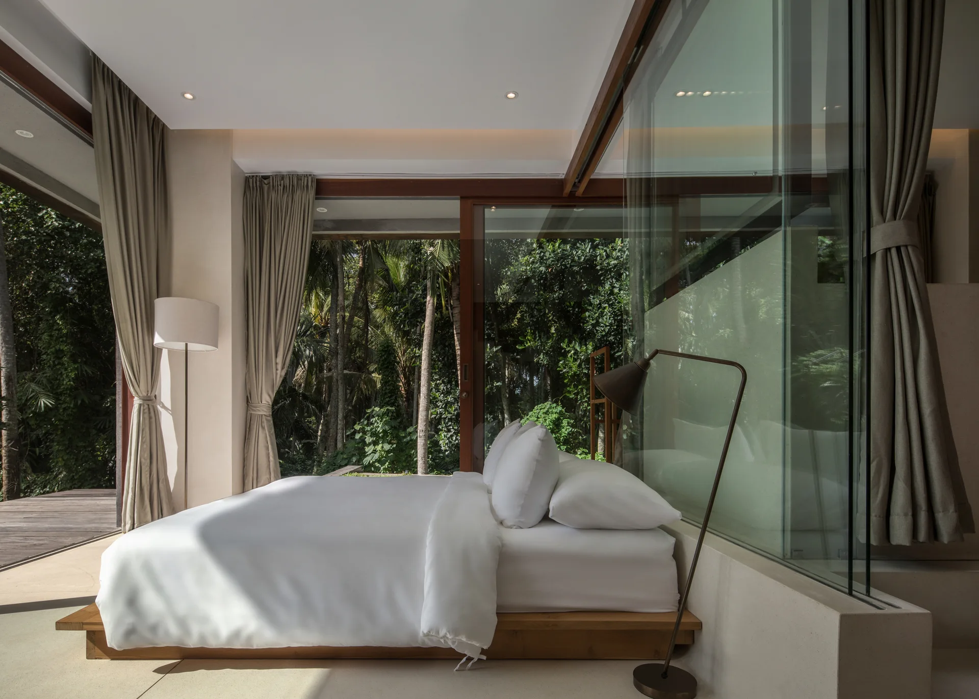 A minimalist bedroom features a low wooden platform bed next to a large glass wall partition and sliding doors opening onto a tropical forest view.