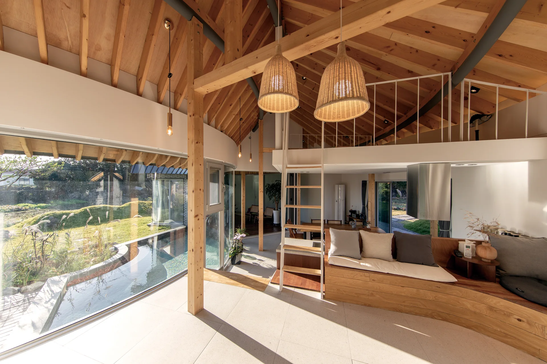 An open living space with a curved glass wall overlooking a garden, featuring exposed timber ceiling beams and a custom wooden bench.