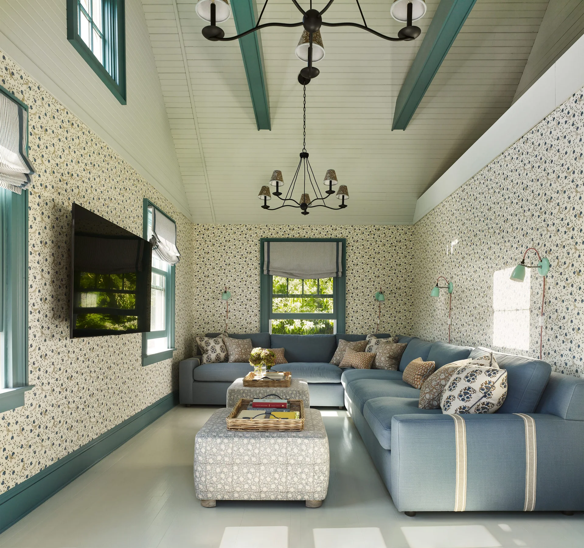 A cottage-style living room features a blue sectional sofa, patterned ottomans, floral wallpaper, and teal-painted window trim and ceiling beams.