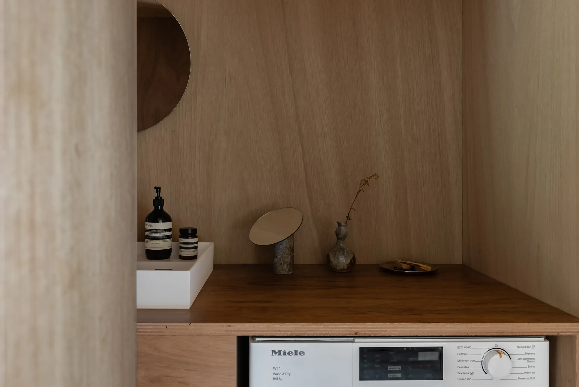 A washing machine is integrated into warm wood cabinetry beneath a countertop with a white basin and a small round mirror.