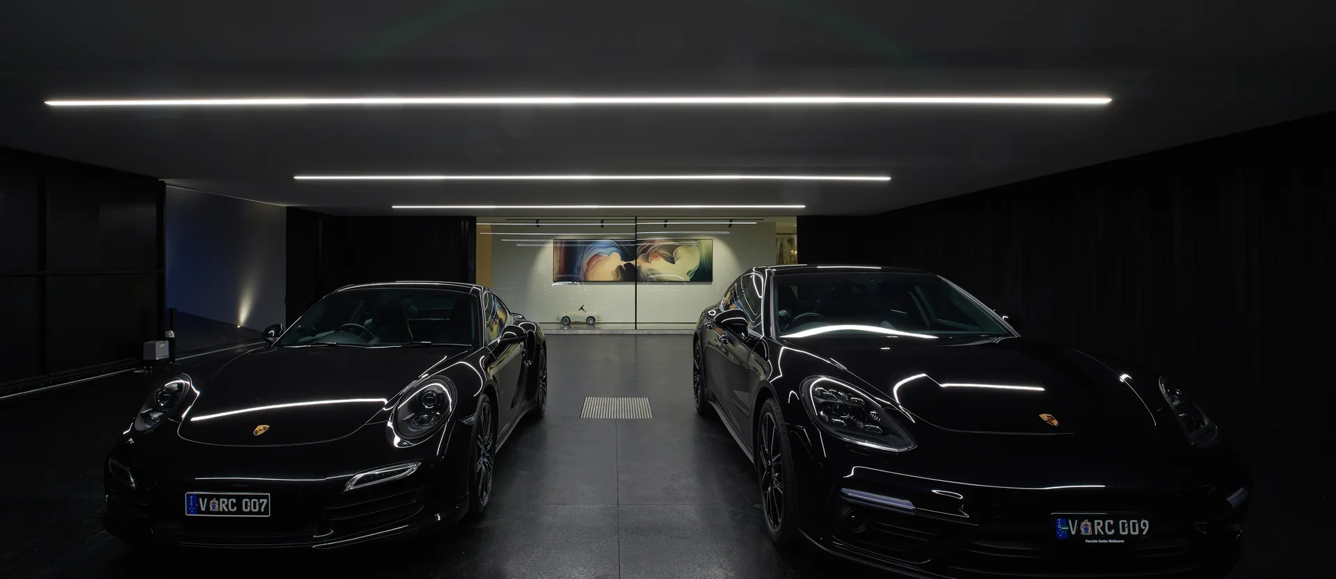 Two black sports cars are parked inside a dark minimalist garage featuring long linear recessed ceiling lights and black vertical wall paneling.