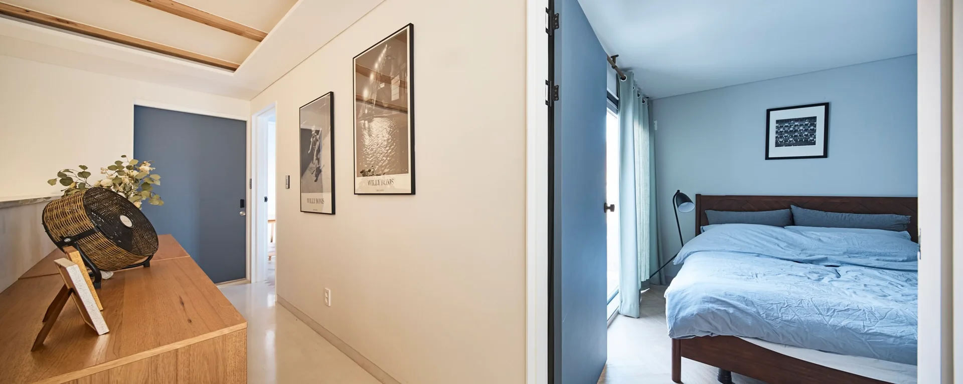 A view from a light beige hallway with framed art into a bedroom featuring dusty blue walls, a wooden bed, and light blue bedding.