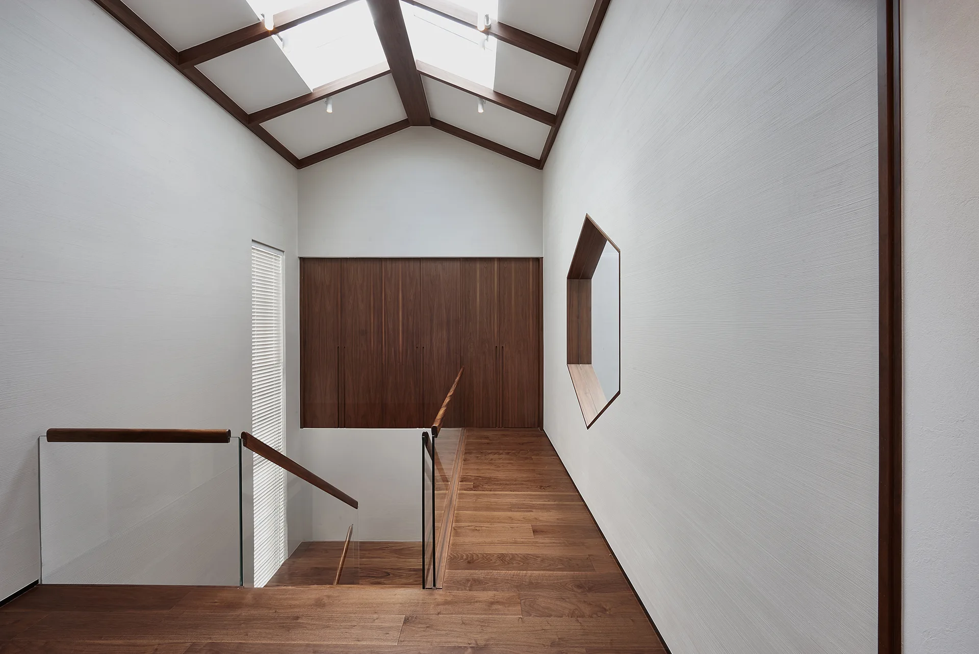 A modern minimalist staircase landing featuring warm wood flooring, a glass railing, and a geometric hexagonal wall niche under skylights.
