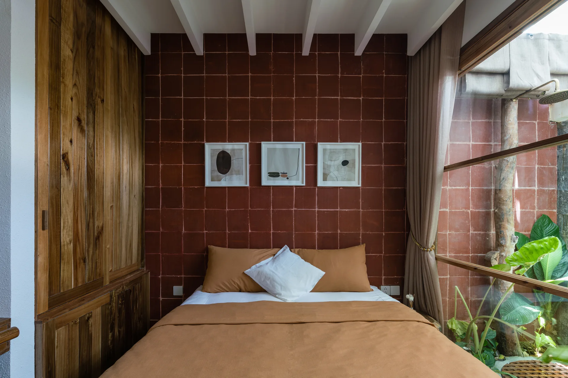 A bed with tan bedding is positioned against a terracotta tile accent wall next to a large window looking onto a small indoor garden.