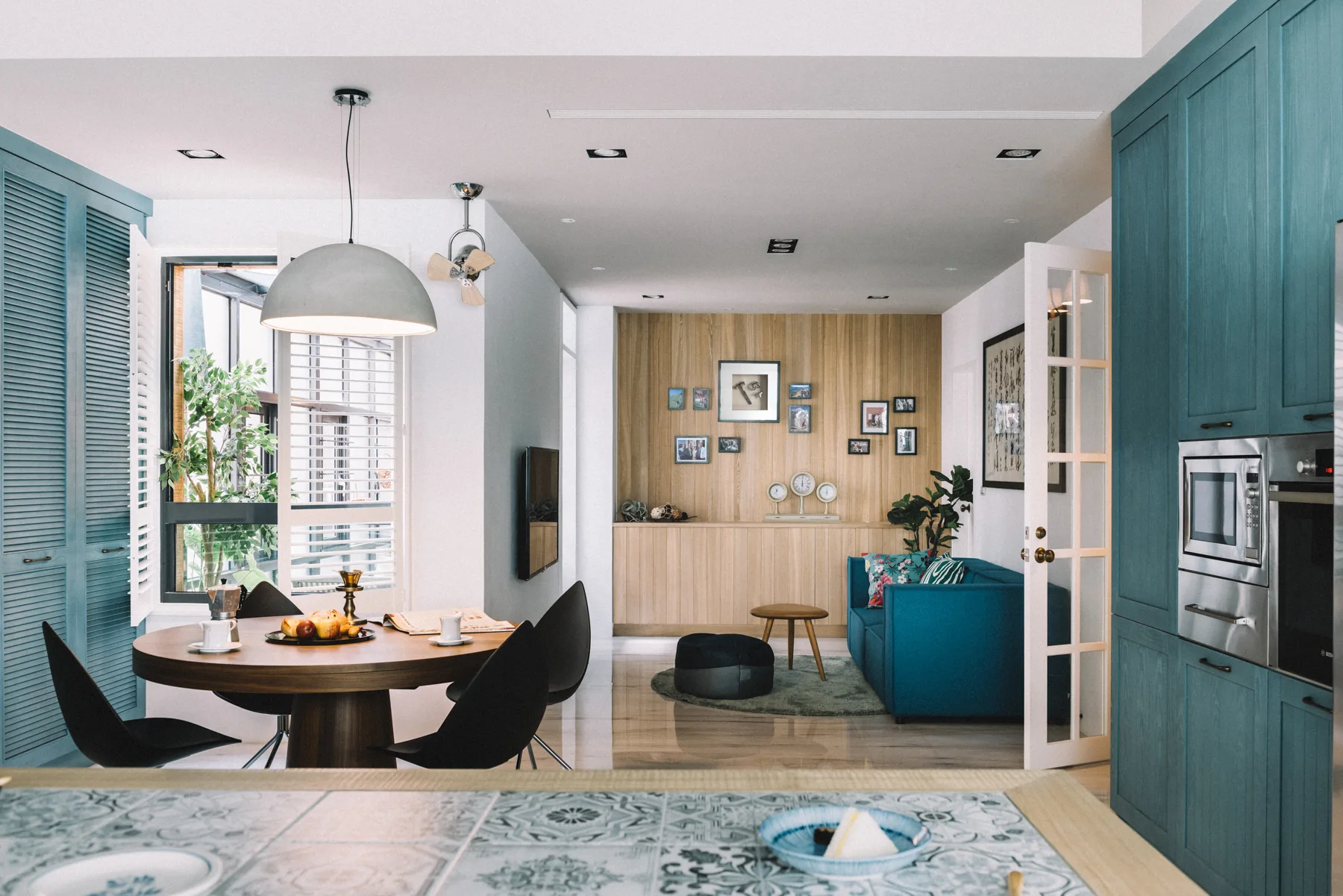 An open-plan residential interior with a round wooden dining table, black chairs, teal kitchen cabinetry, and a wood-paneled feature wall.