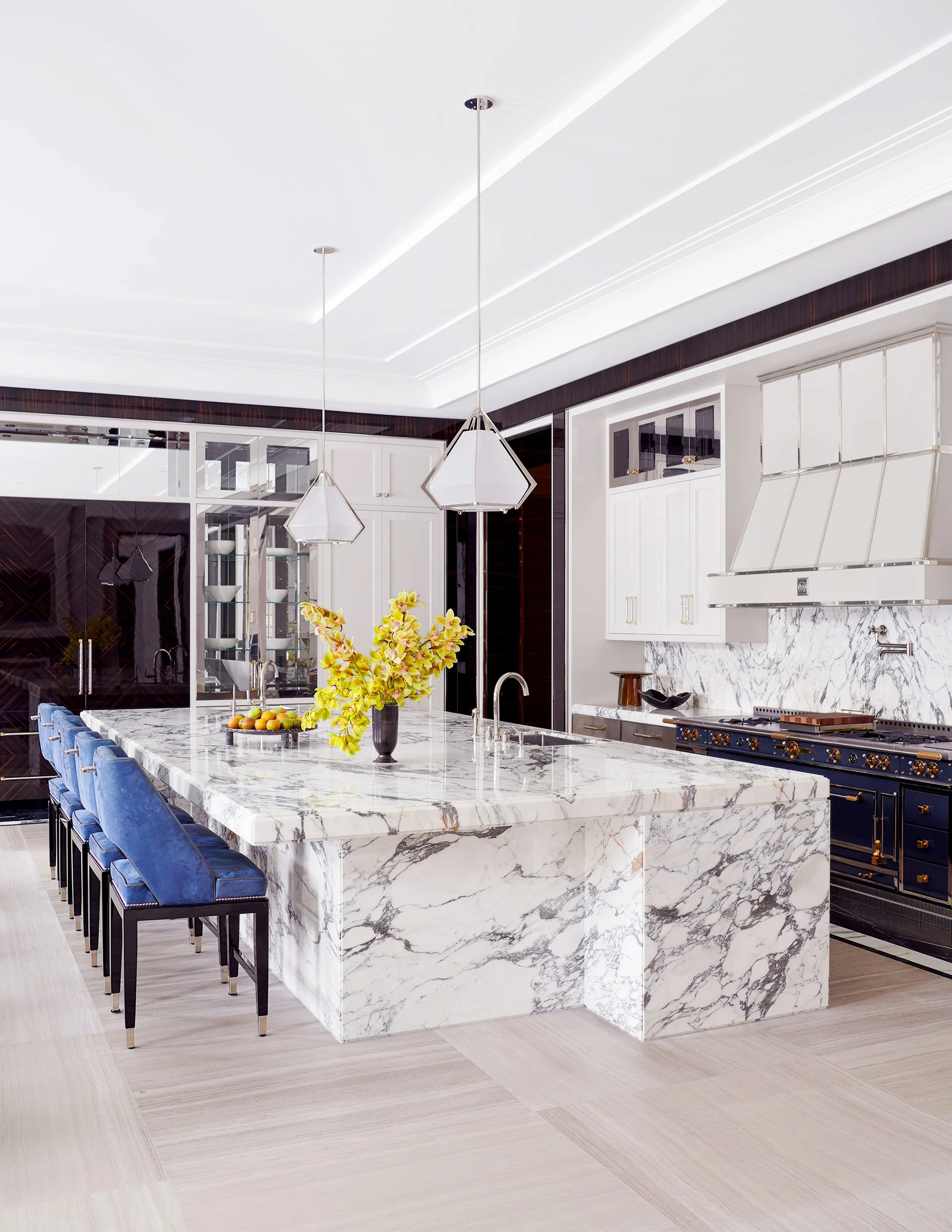 A large marble-topped kitchen island with royal blue bar stools stands in a modern kitchen next to a navy blue professional range.
