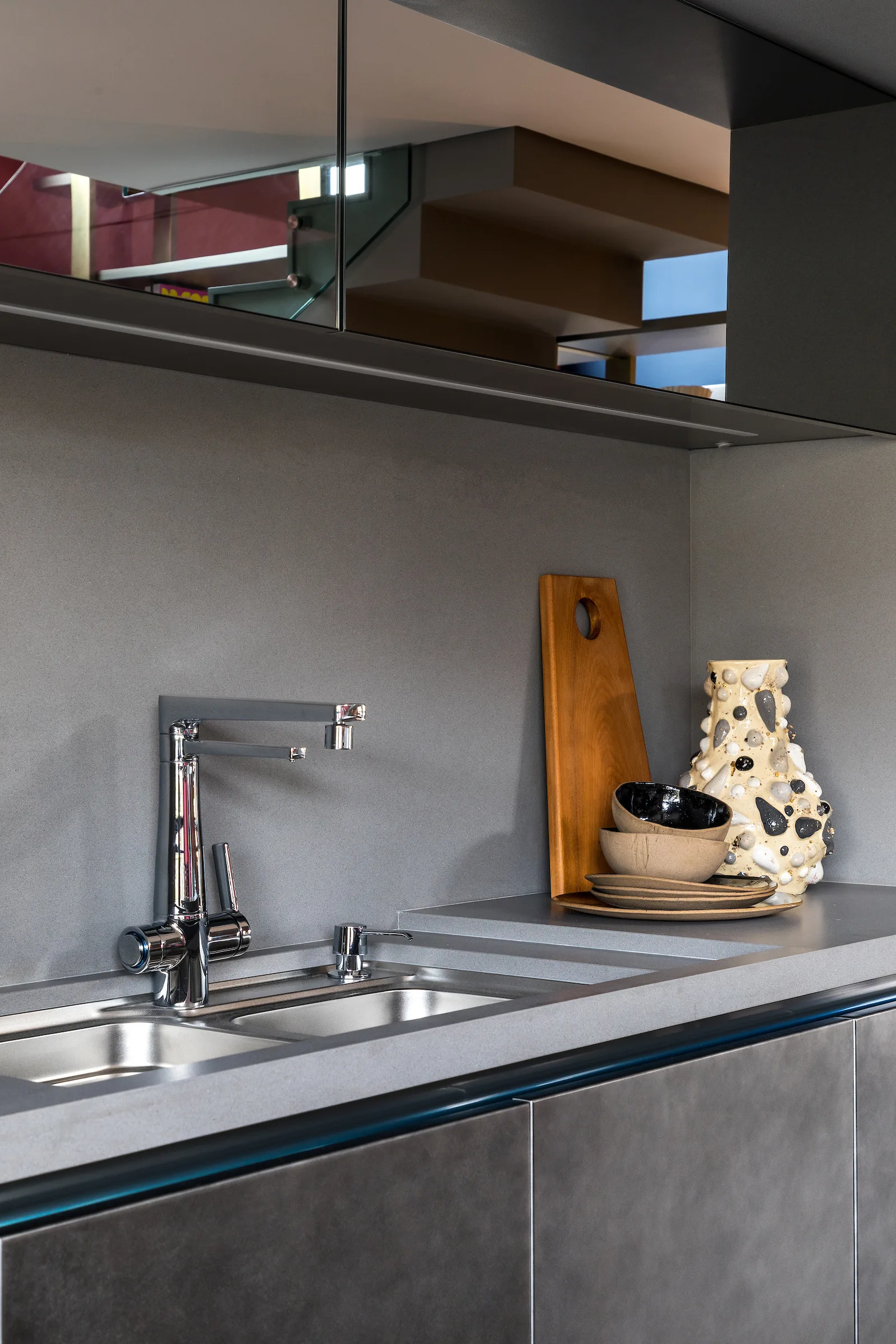 A close-up of a grey kitchen countertop with a chrome faucet, integrated undermount sink, wooden cutting board, and textured ceramic vase.
