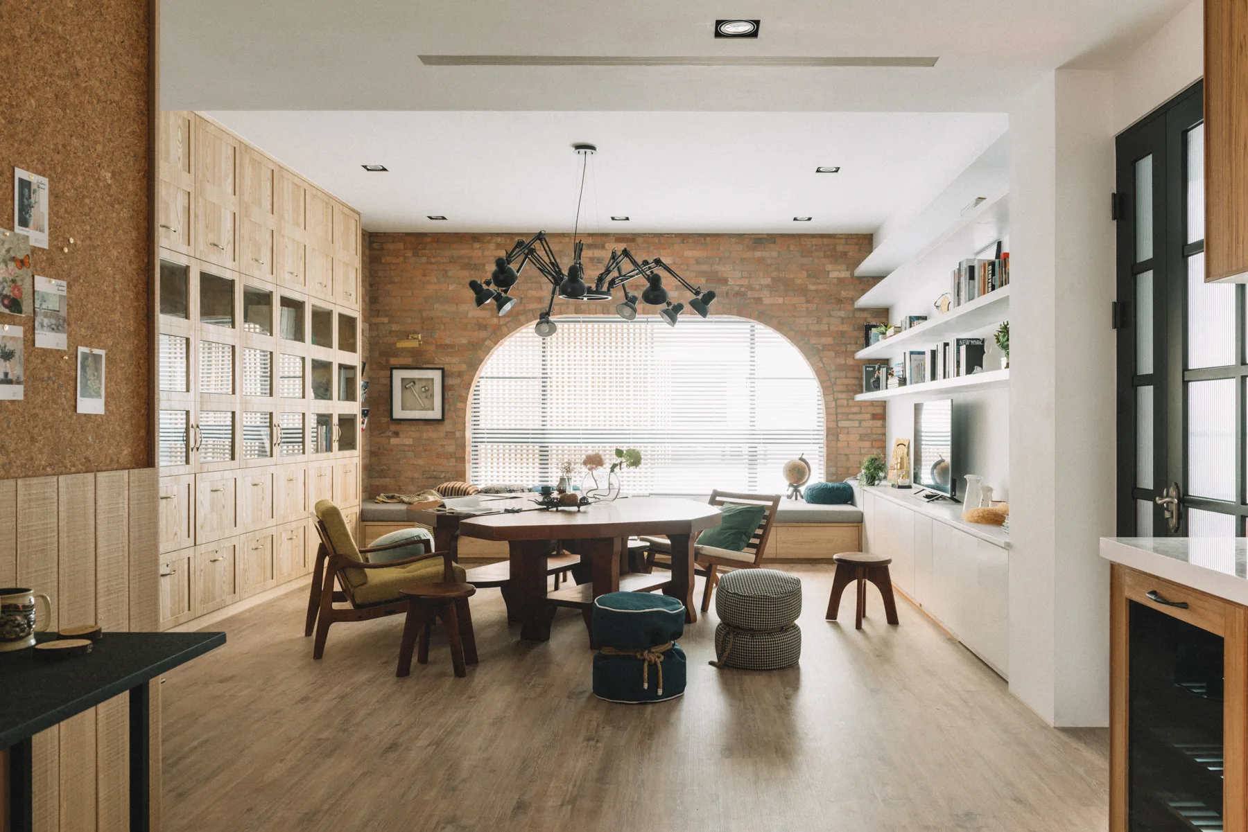 An eclectic living room features a central wooden dining table, a large arched window set in an exposed brick wall, and extensive built-in wooden cabinetry.