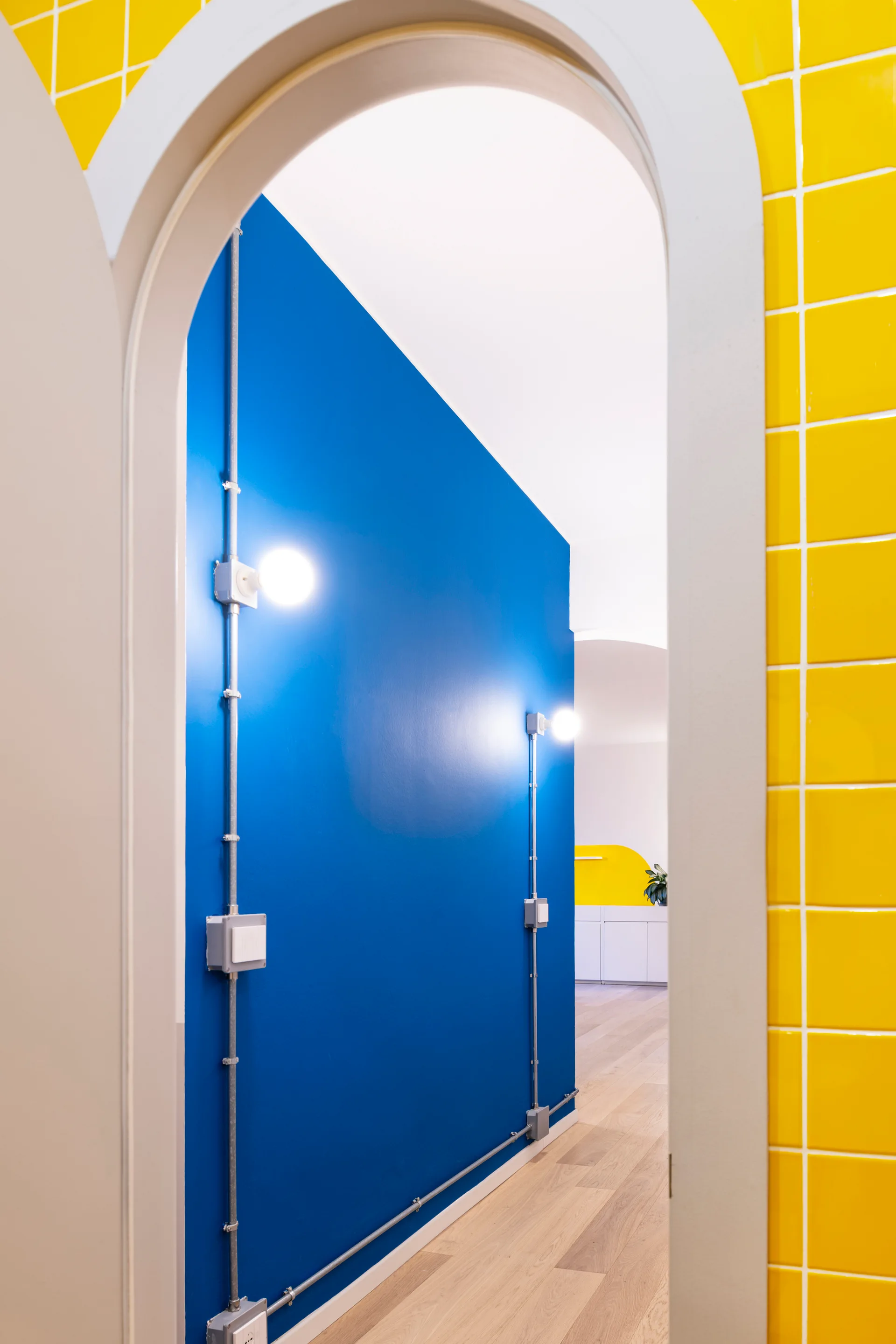 An archway frames a hallway featuring a bold blue wall with exposed metal conduits and bare bulb lights, adjacent to a yellow tiled wall.