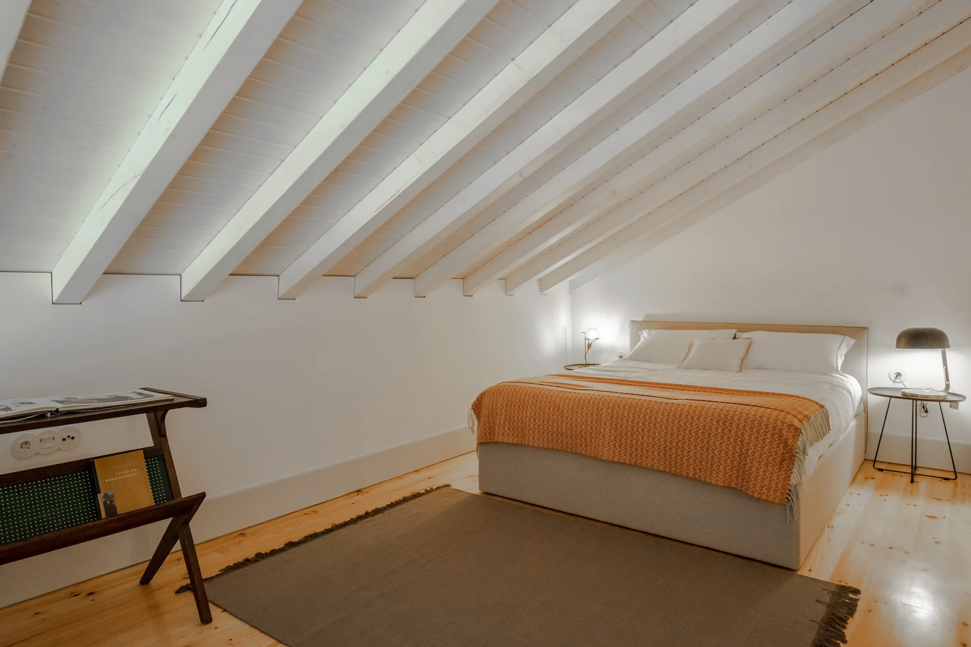 A minimalist attic bedroom features a low bed with an orange throw blanket under a sloped white ceiling with exposed painted wooden beams.