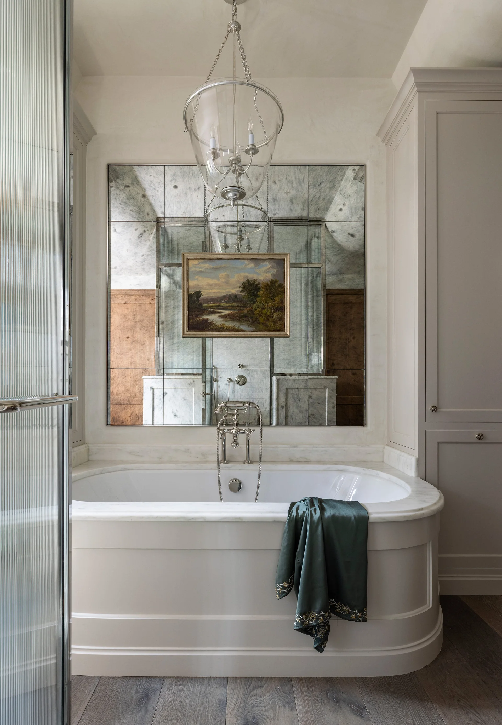 A white soaking tub with a marble ledge sits beneath an antiqued mirrored wall featuring a hung landscape painting and a glass lantern light fixture.