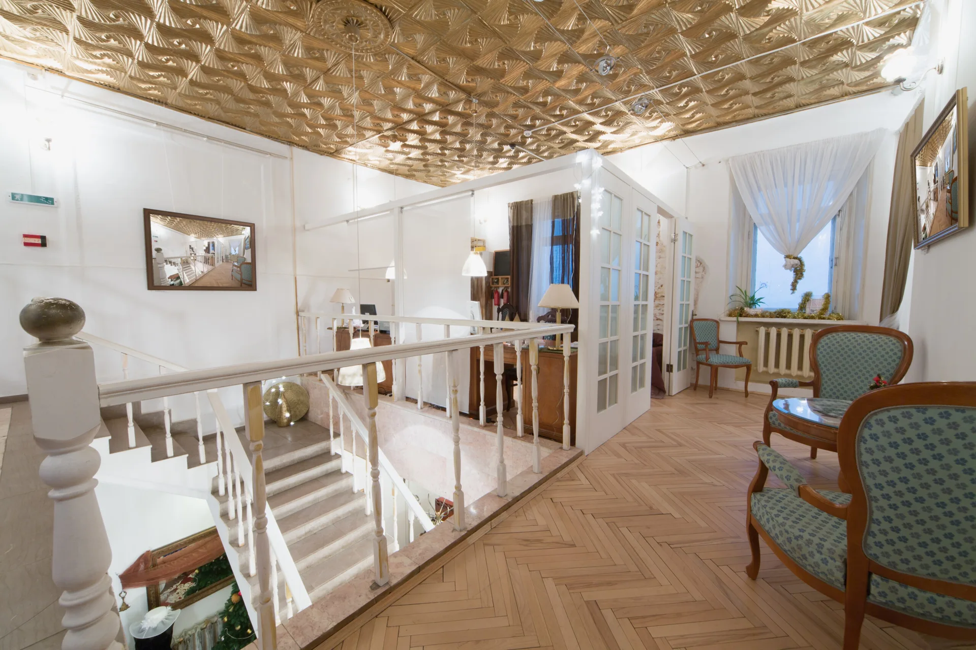 A staircase landing features an ornate gold geometric ceiling, light wood herringbone flooring, white spindle railings, and patterned armchairs.