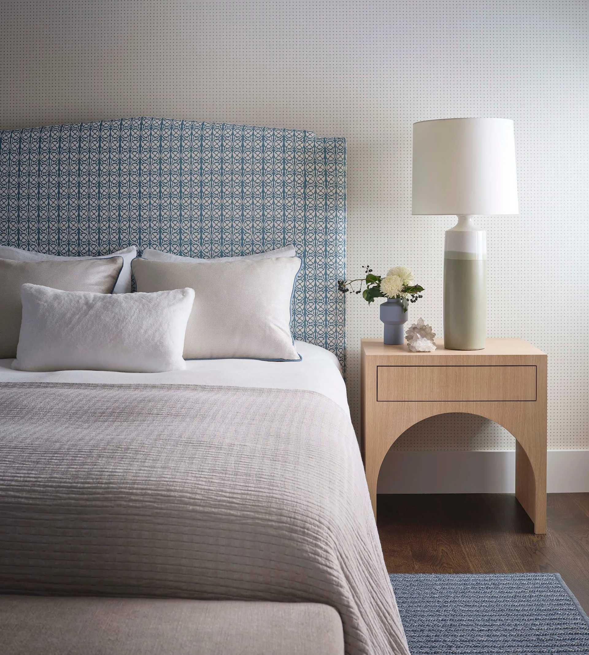 A bedroom features an upholstered blue patterned headboard against dotted wallpaper, paired with a light wood arched nightstand and ceramic table lamp.