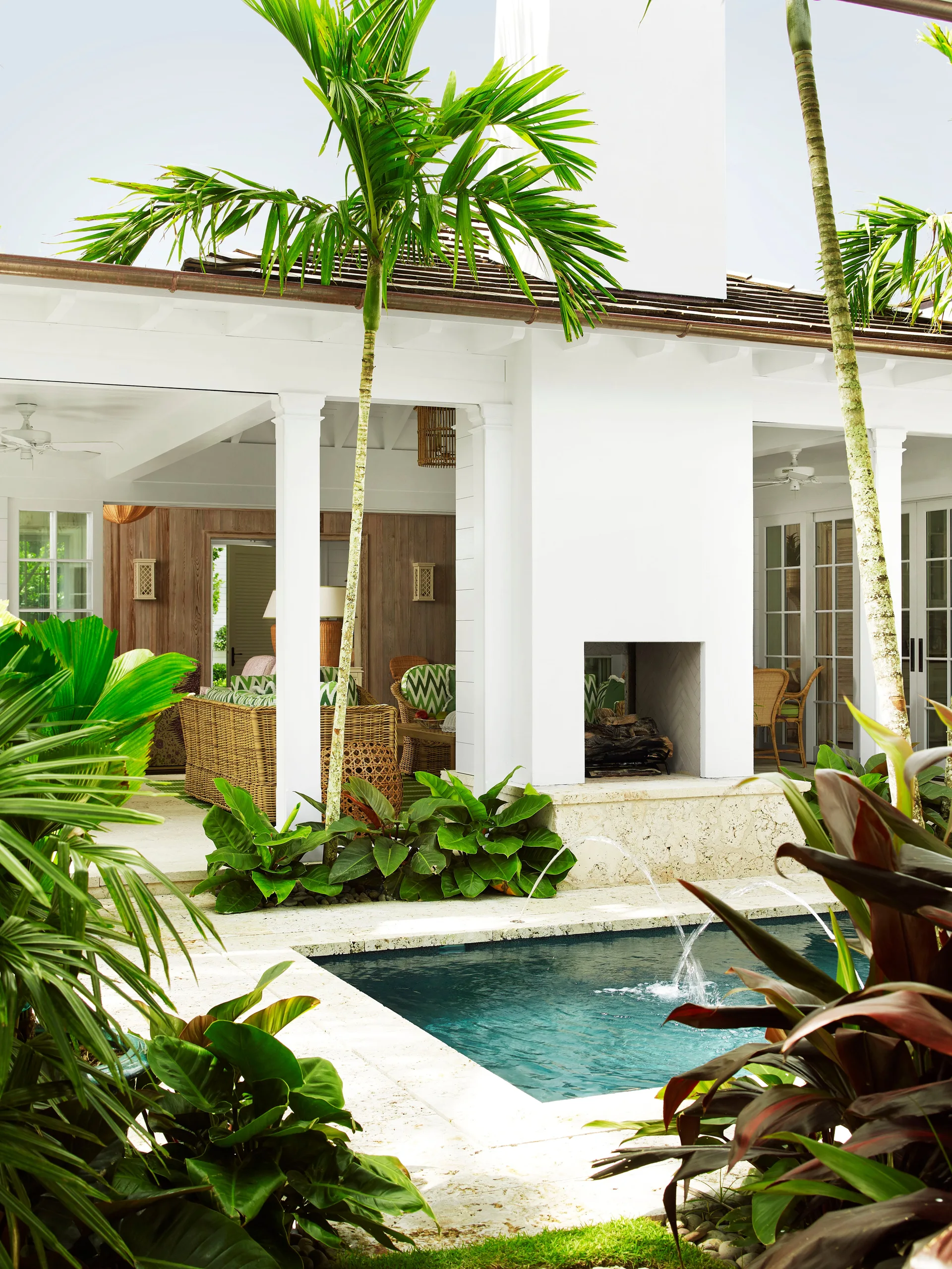 A covered patio with a white fireplace and wicker furniture next to a swimming pool with water fountains surrounded by tropical palm trees.