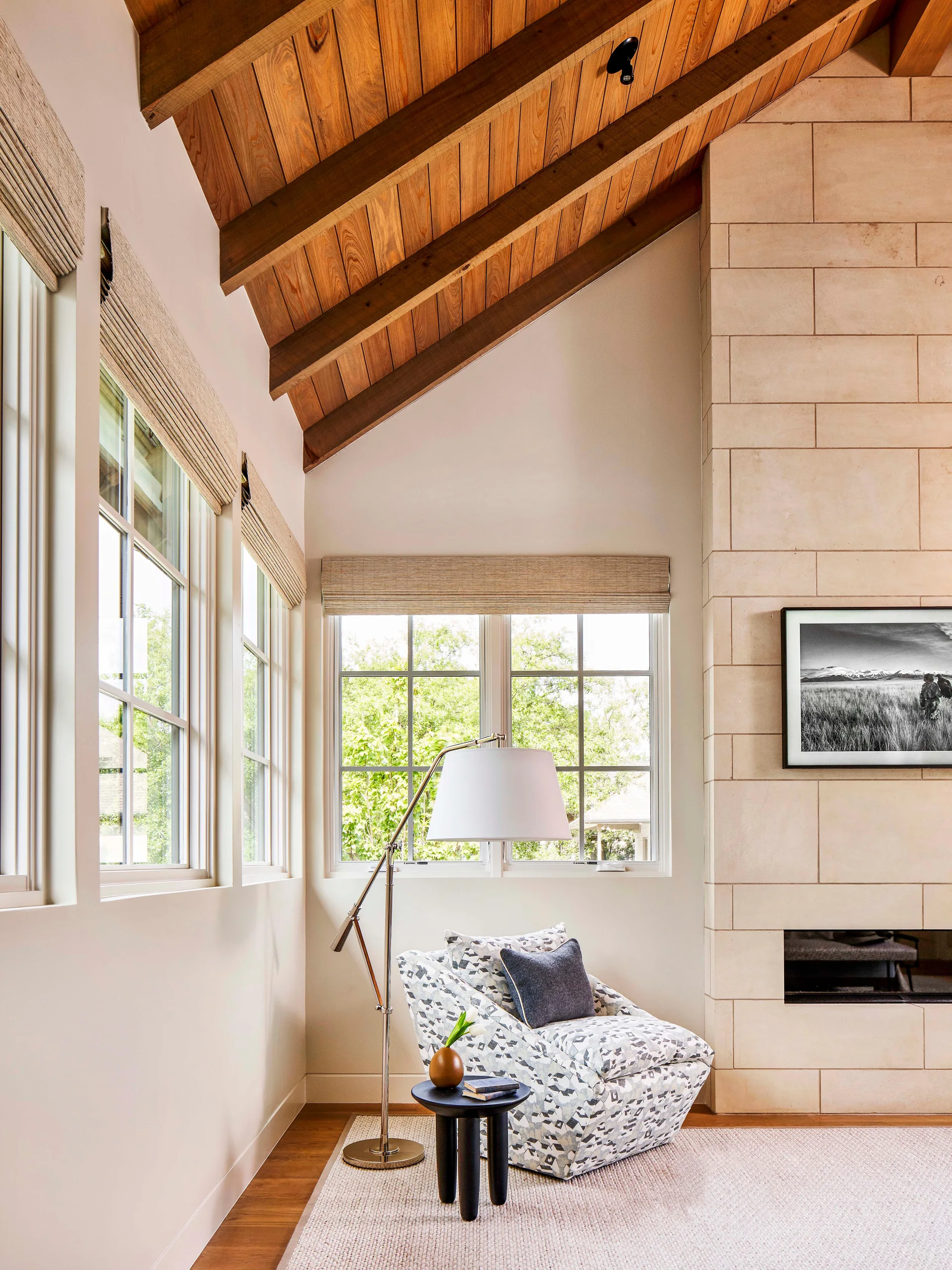 A patterned armchair, a floor lamp, and a small black side table sit beside a limestone fireplace beneath a vaulted wood ceiling.