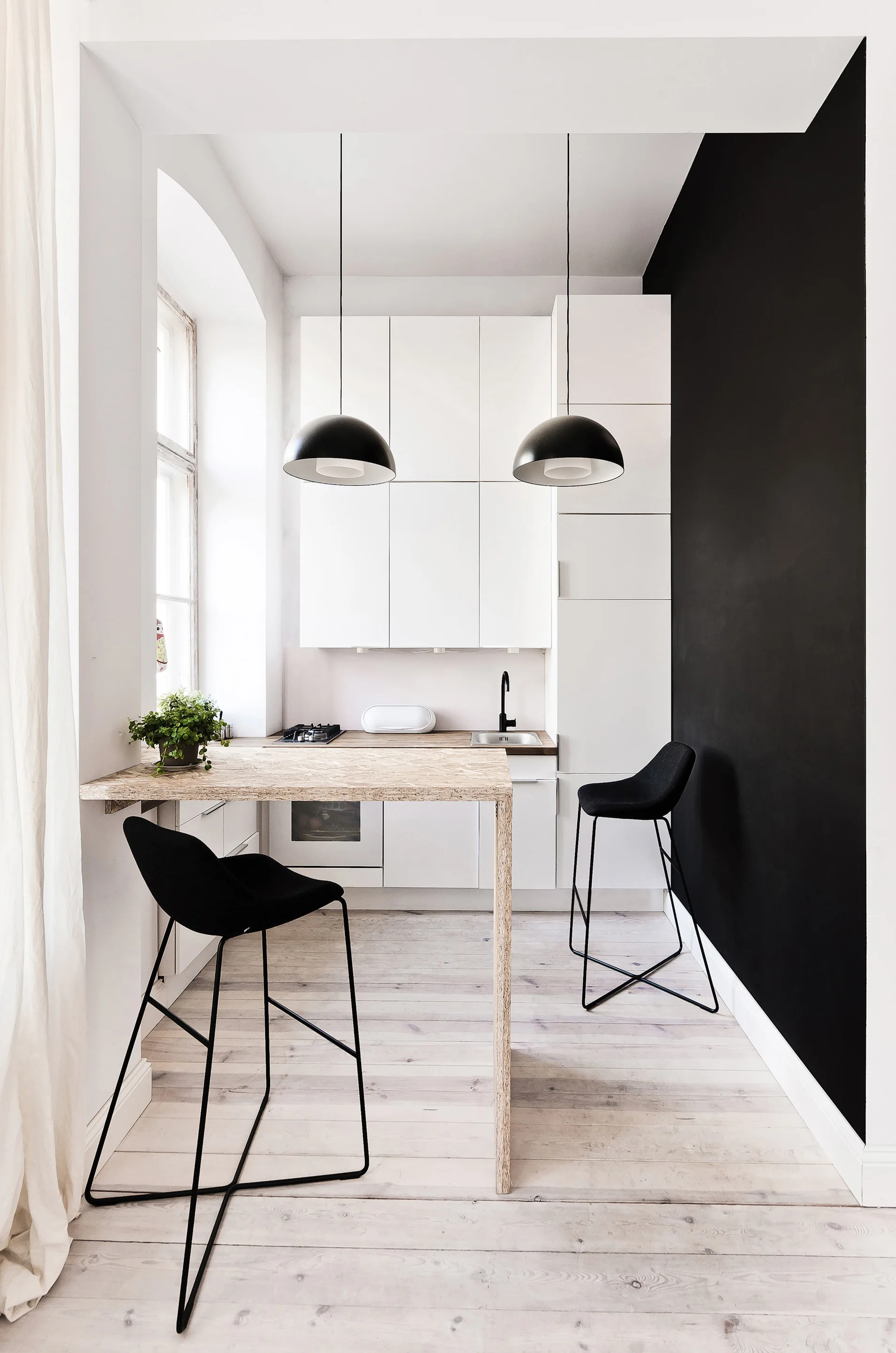 A minimalist kitchen features white flat-front cabinets, a black accent wall, two sleek black stools, and a wood-textured breakfast bar.