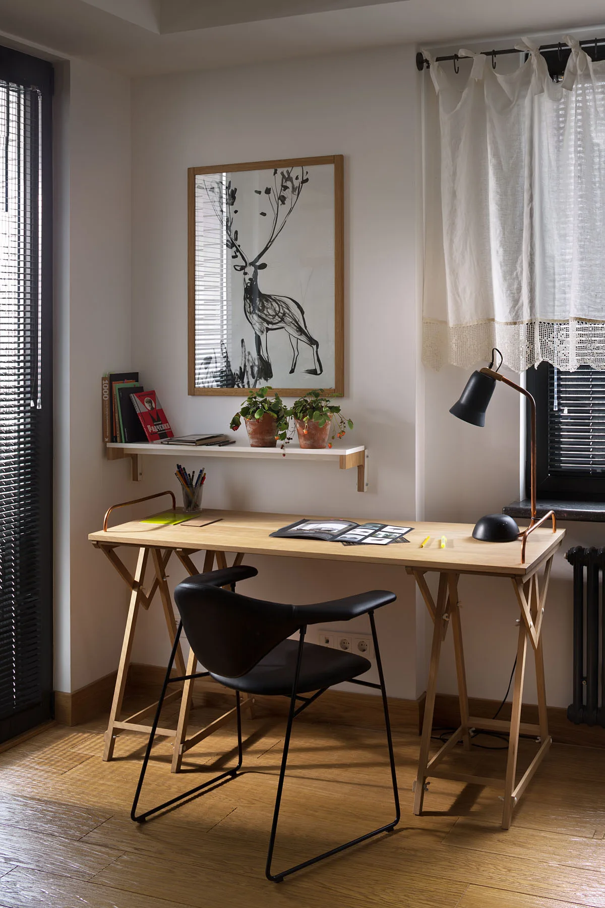 A light wood trestle desk with a black desk lamp and chair sits on wood flooring below a white wall shelf with framed artwork and potted plants.