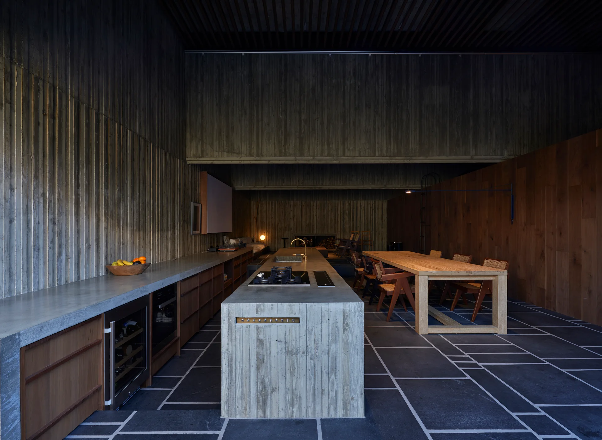 A minimalist kitchen features a raw concrete island, dark stone-tiled floors, ribbed concrete walls, and a large wooden dining table with matching chairs.