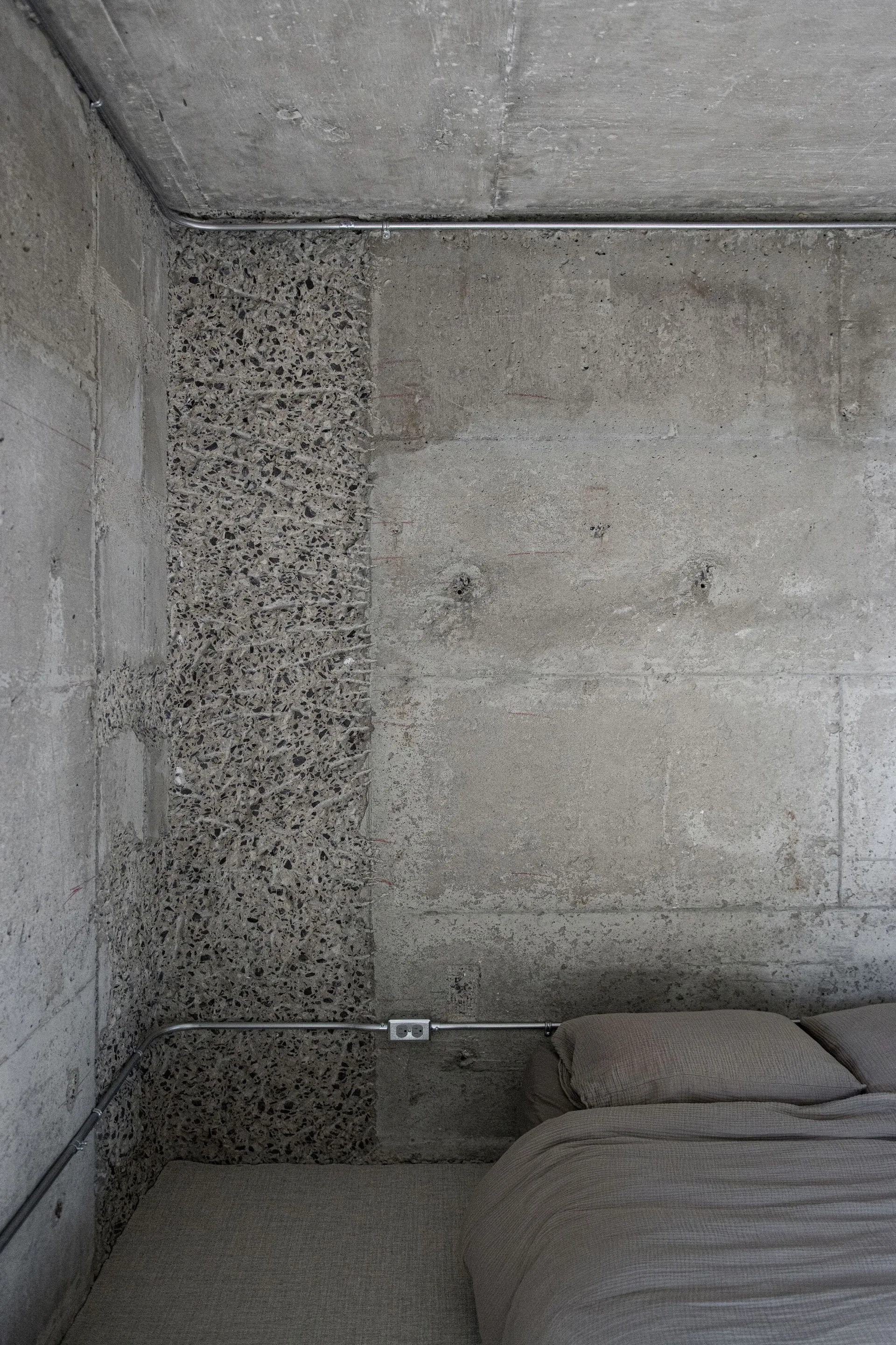 A bedroom corner features raw concrete walls with a textured aggregate strip, an exposed metal conduit, and a bed with gray linens.