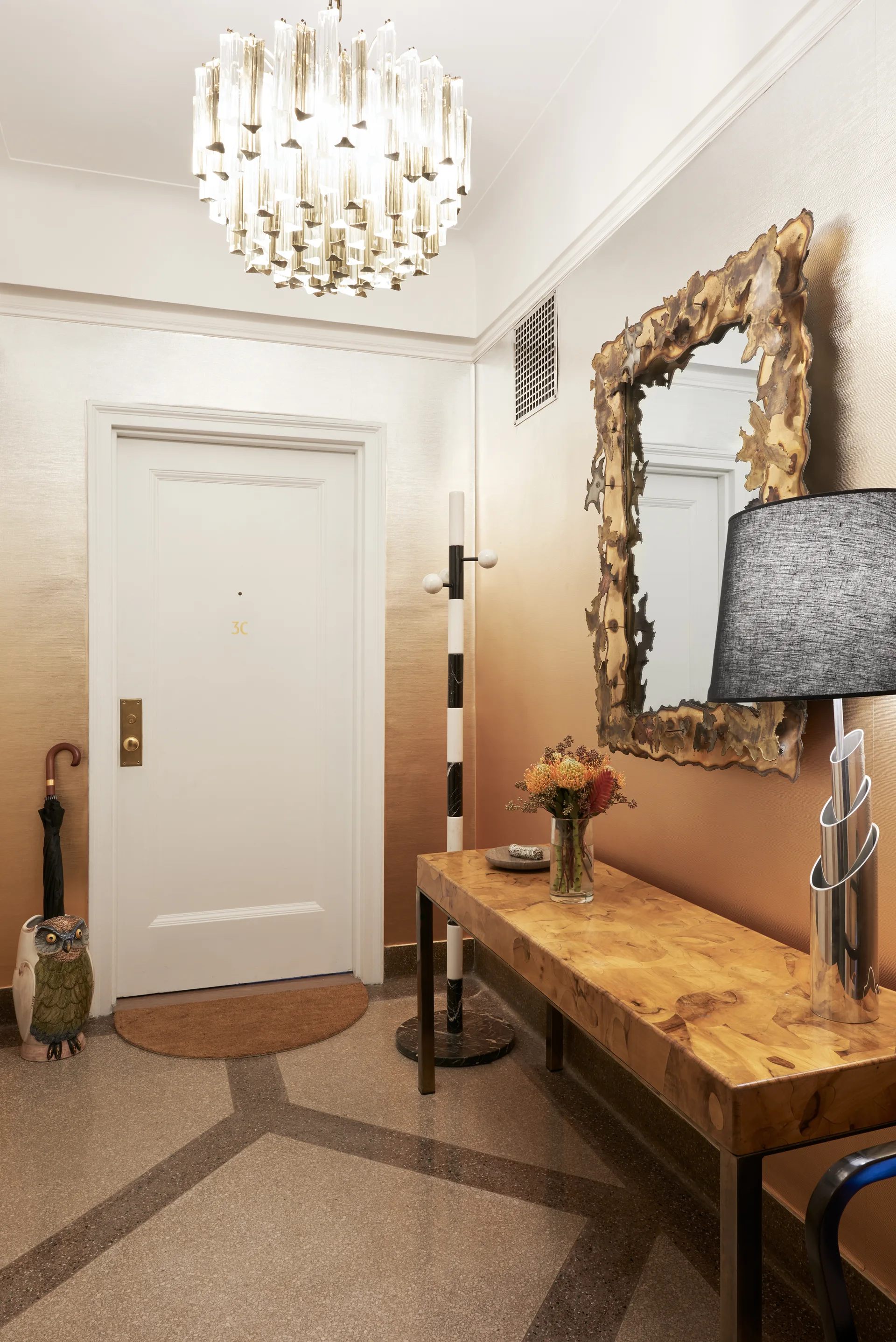 An entryway features gold gradient wallpaper, a burl wood console table, a sculptural metallic mirror, and a tiered glass chandelier.