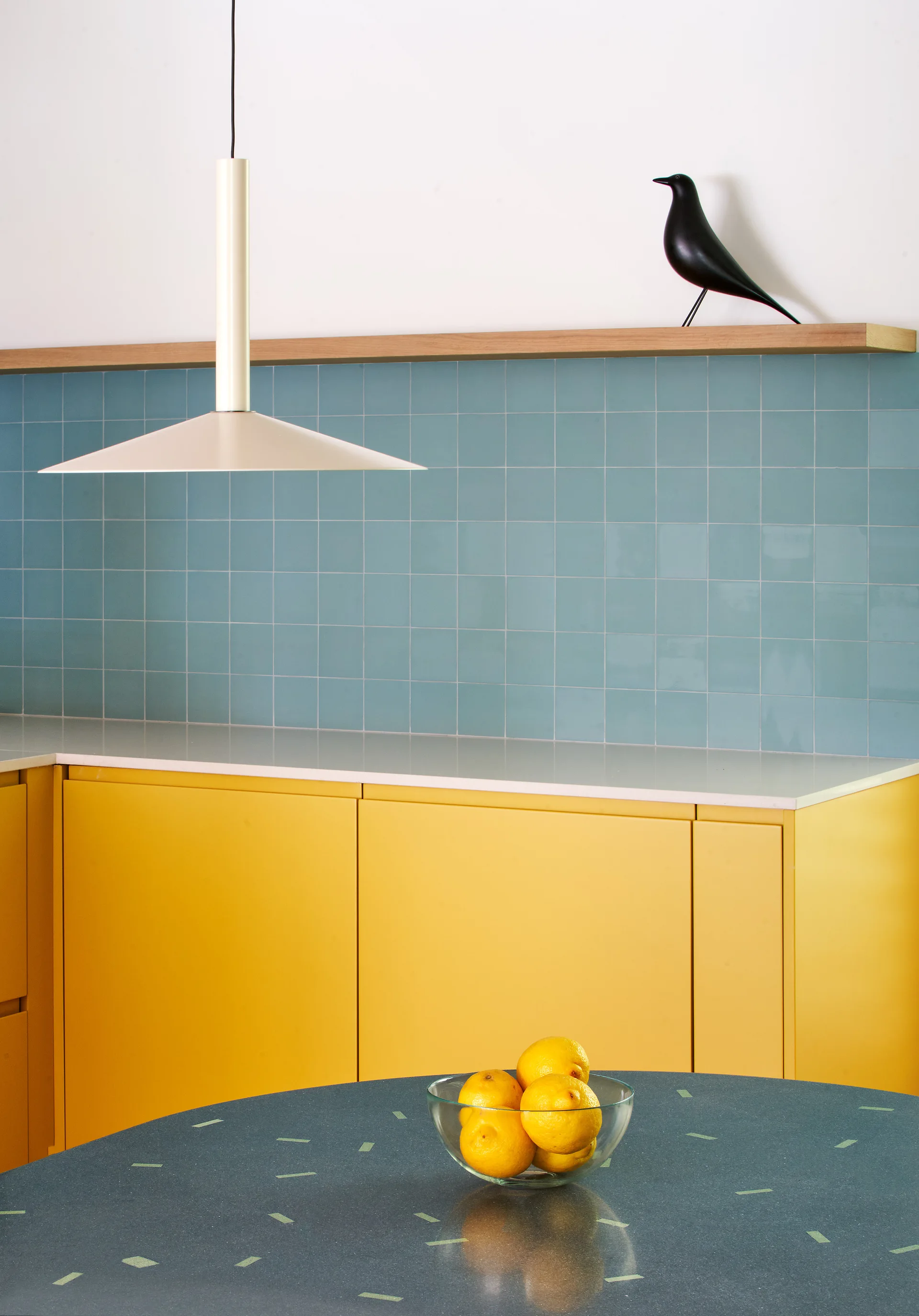 A modern kitchen view showcasing yellow cabinetry, a glossy blue tile backsplash, a floating wooden shelf, and a white pendant light.