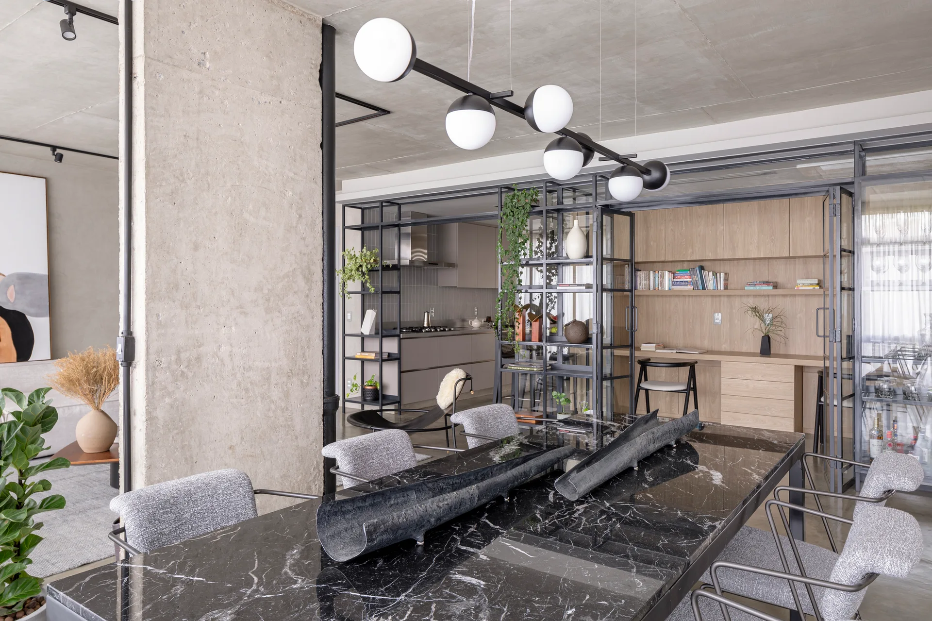 A modern dining room features a black marble table with grey chairs beneath a globe chandelier, with a glass partition and concrete pillar nearby.