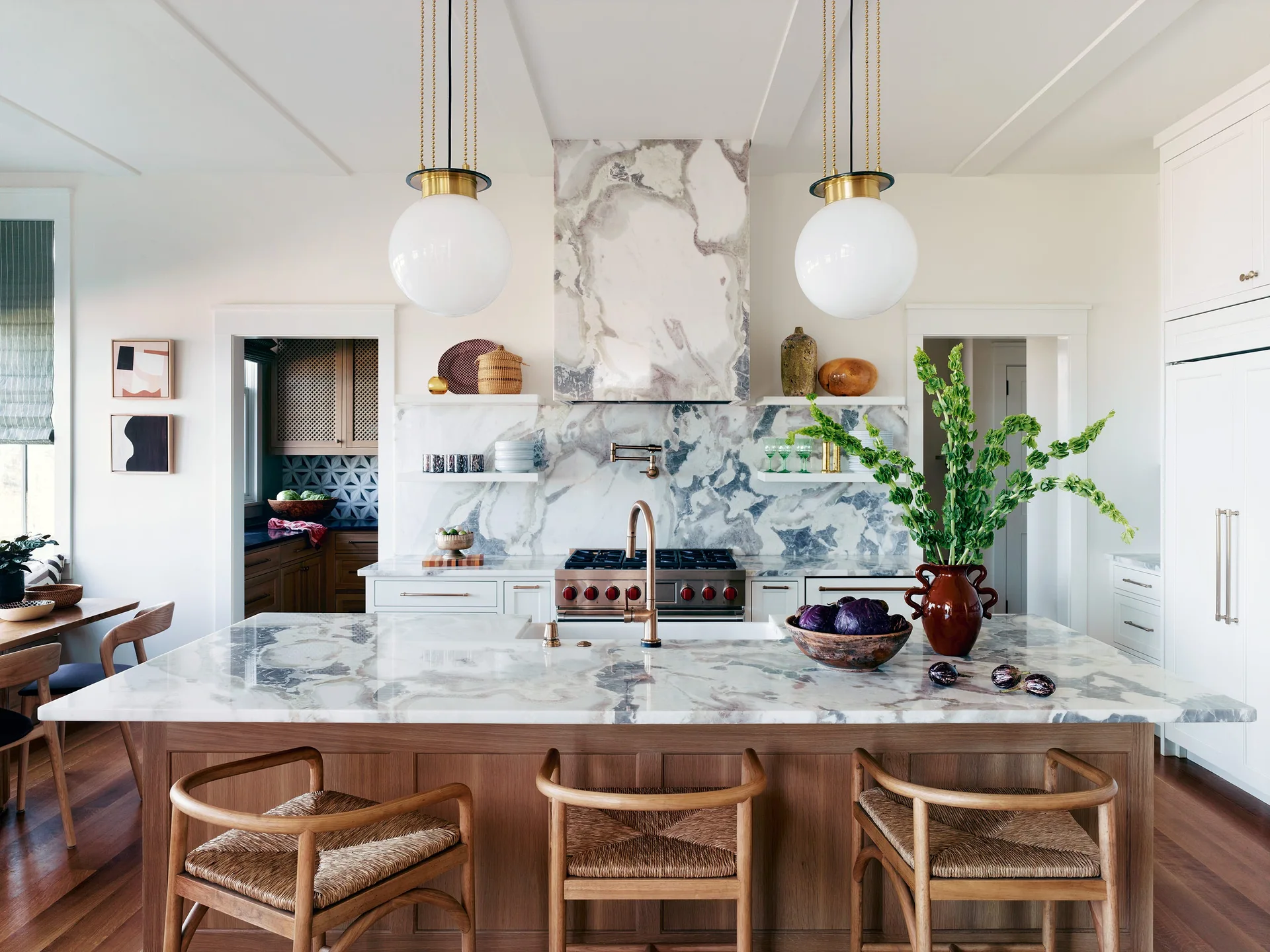 A bright kitchen features a wooden central island with a white marble countertop, matching slab backsplash, three woven counter stools, and globe pendants.