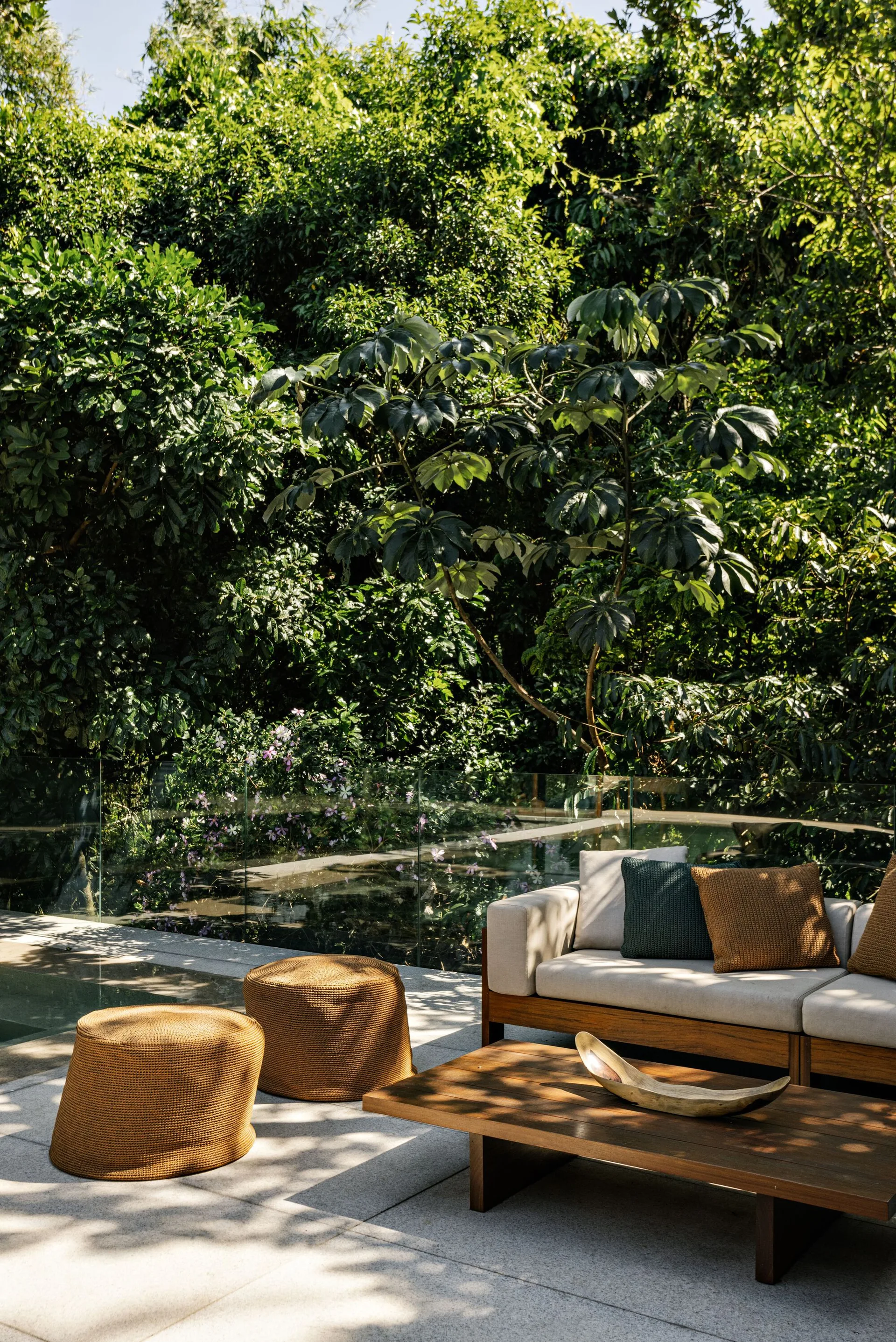 A modern wood-framed outdoor sofa with beige cushions sits on a stone patio alongside two woven poufs and a low wood coffee table.