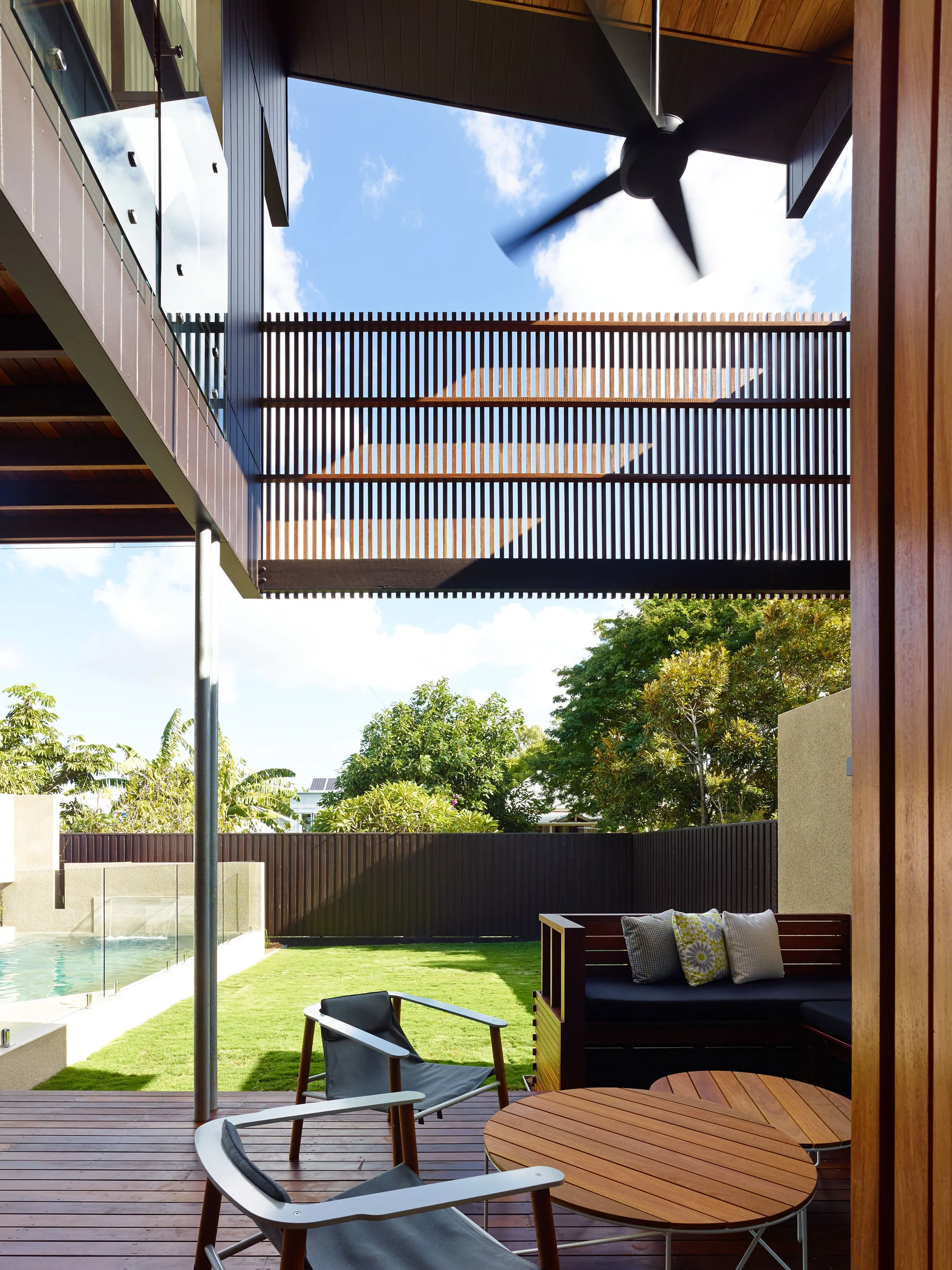A covered wooden deck features modern outdoor lounge furniture overlooking a grassy yard and swimming pool under a bright blue sky.