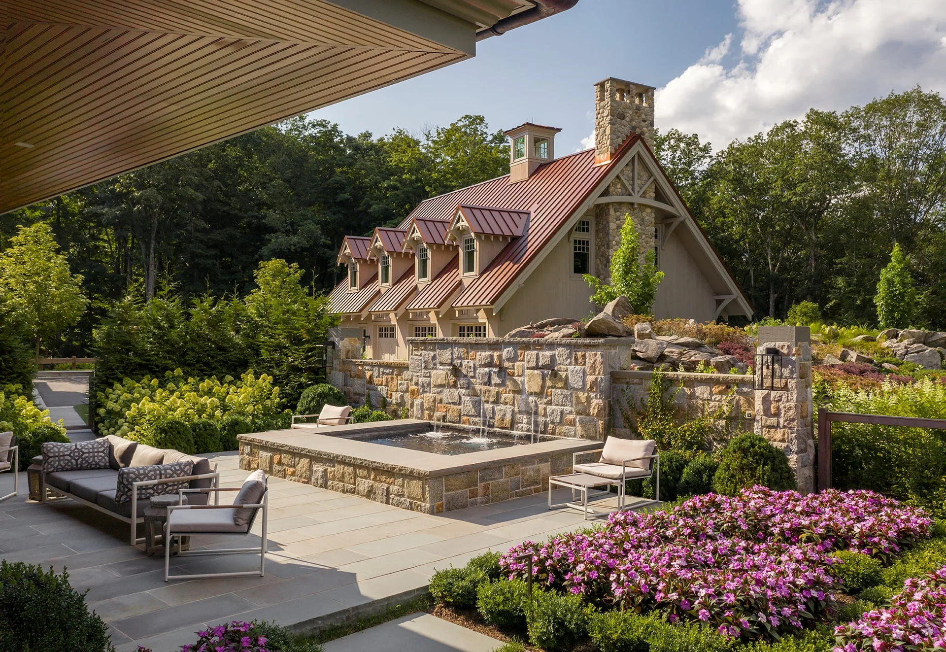 A stone patio with modern grey outdoor sofas surrounds a built-in stone fountain, with a dormered house and colorful gardens in the background.