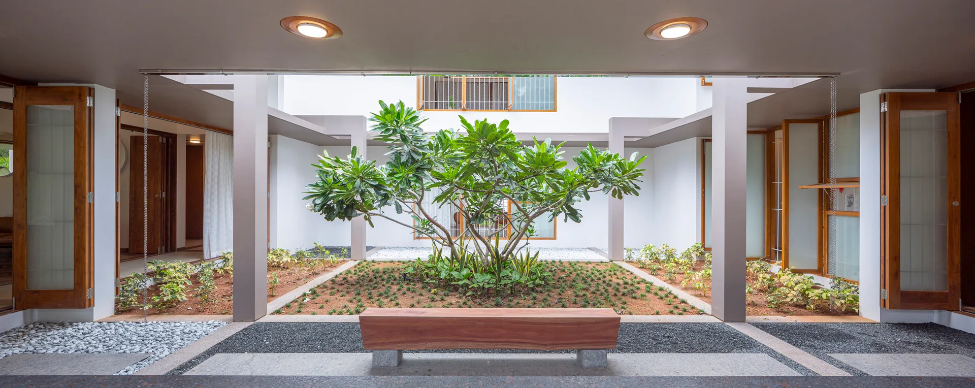 A symmetrical outdoor courtyard featuring a central garden bed with a green tree, surrounded by a covered walkway with columns and a wooden bench.
