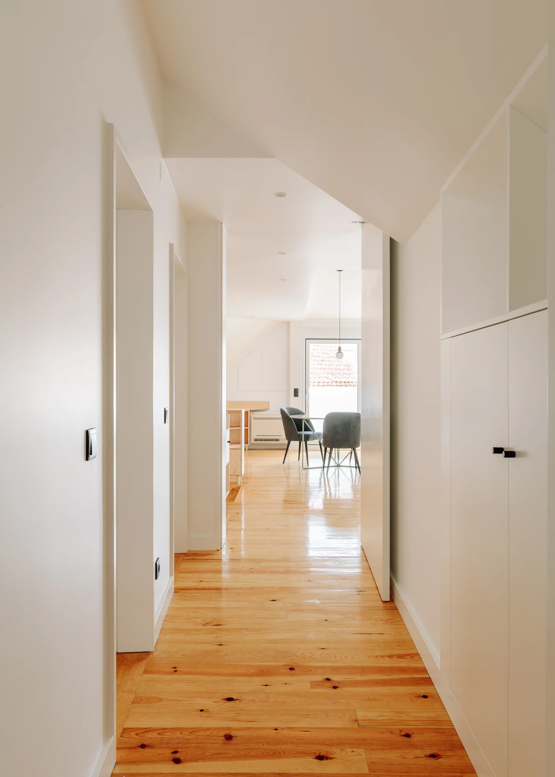 A minimalist hallway with glossy wood plank flooring and white walls features built-in cabinets and a view of a dining area.