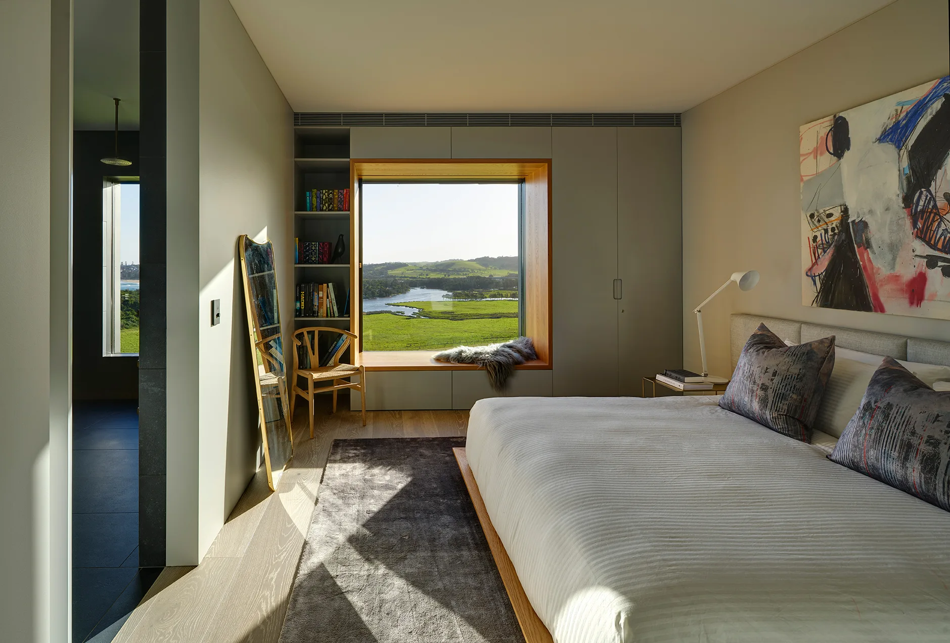 A modern bedroom features a deep wood-framed window seat with green landscape views, integrated grey closet doors, and a low-profile bed.