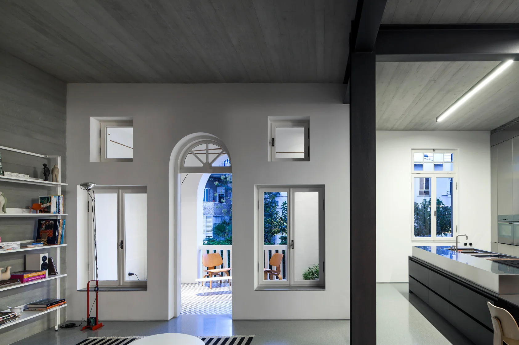 A modern open-concept space featuring a concrete ceiling, white walls with an arched balcony doorway, and a dark grey kitchen island with a metal countertop.