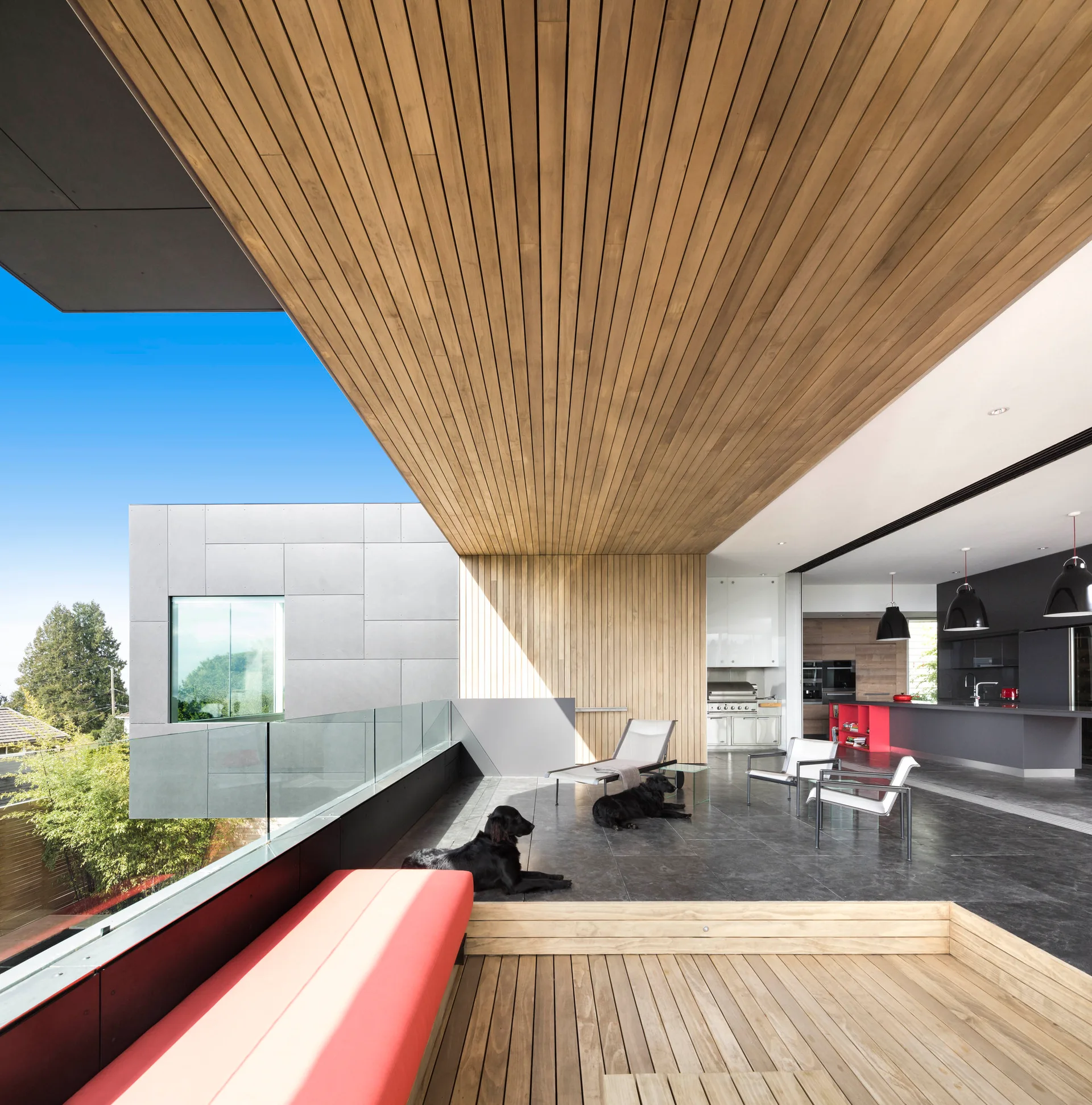 A modern covered terrace featuring dark tile floors, a warm wood-slat ceiling, and a frameless glass railing overlooks a bright outdoor view.
