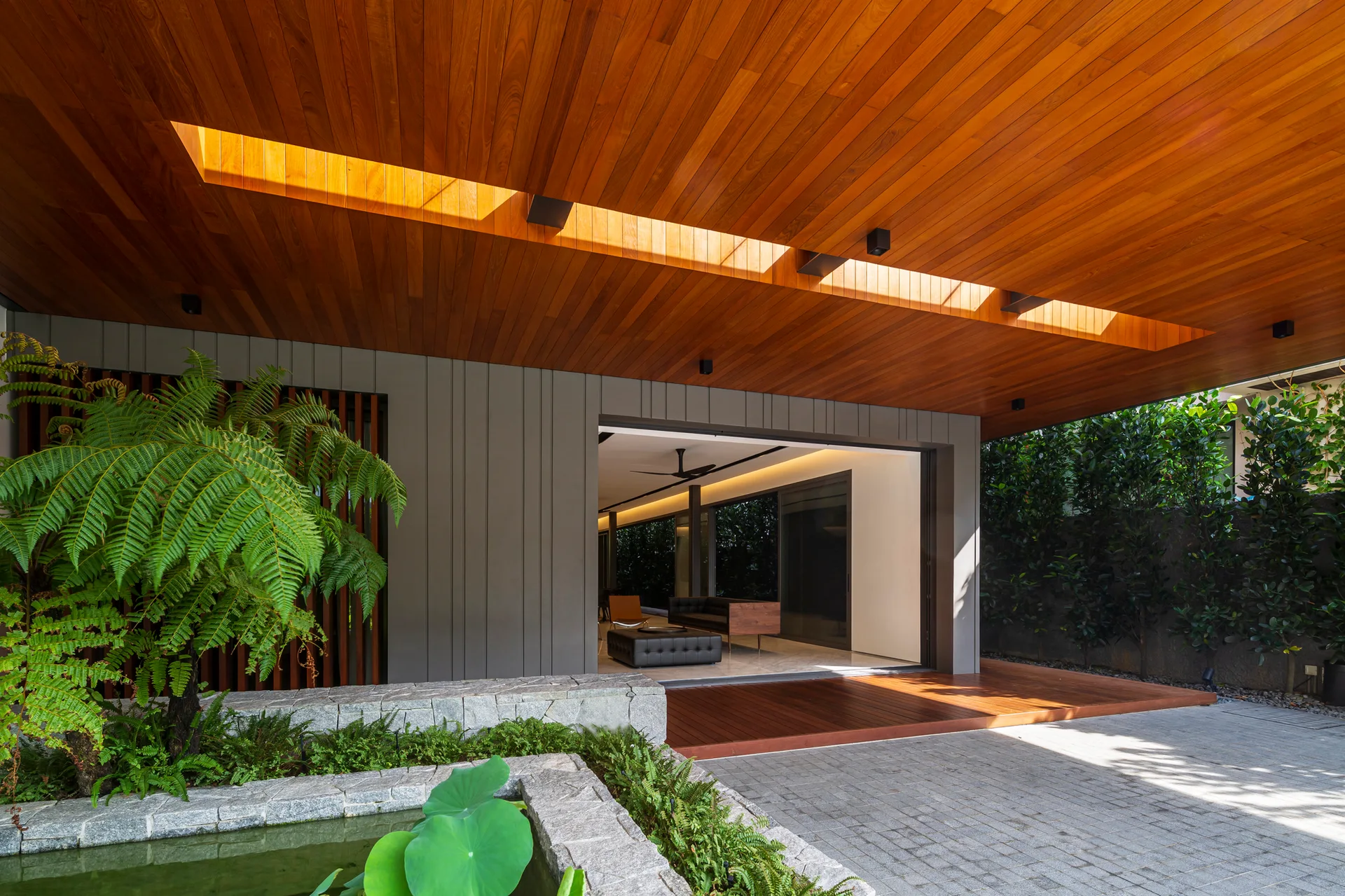 A covered modern patio features a planked wood ceiling with a rectangular skylight, a stone water feature, and grey pavers.